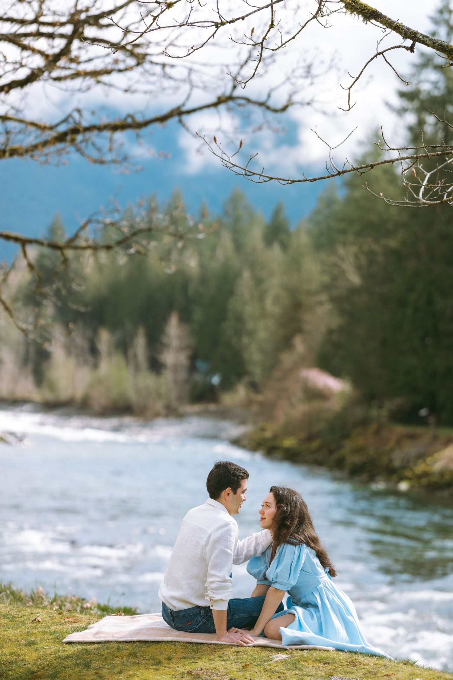 A couple sits on a blanket beside a rushing river surrounded by evergreen trees on a spring afternoon in mountains.