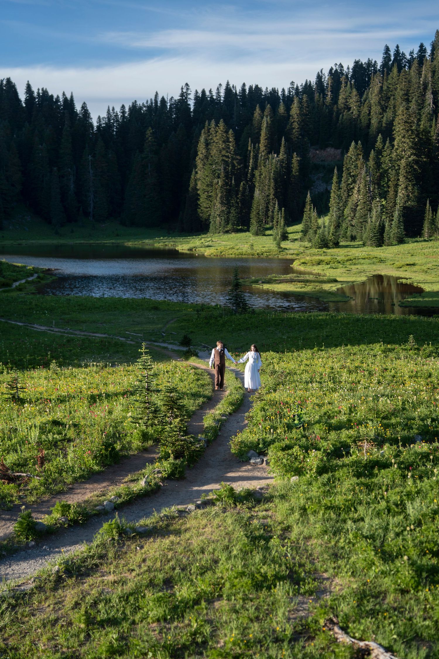 Hikers walk along a scenic mountain trail through alpine meadows beside a serene lake surrounded by evergreen forest.