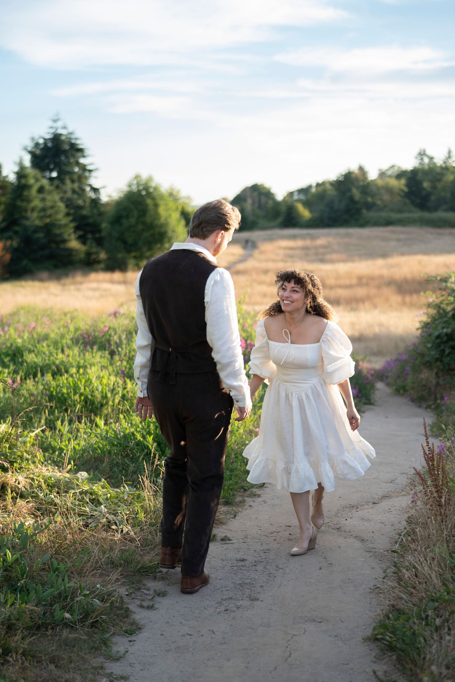 A heartwarming scene shows two figures walking together on a winding dirt path through a summer meadow at sunset.