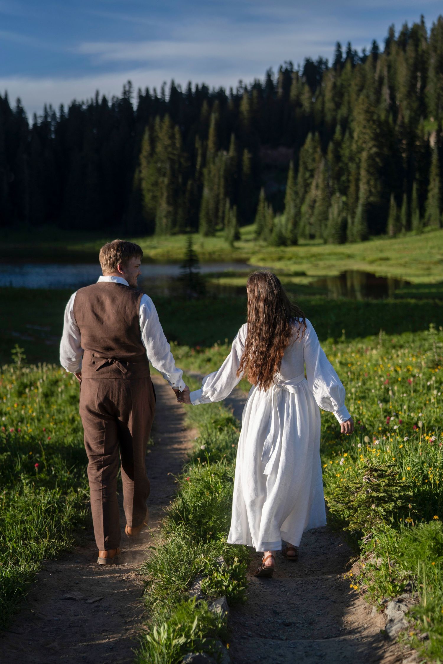 Two people walk hand in hand along a mountain trail through wildflowers and evergreen trees on a sunny summer day.