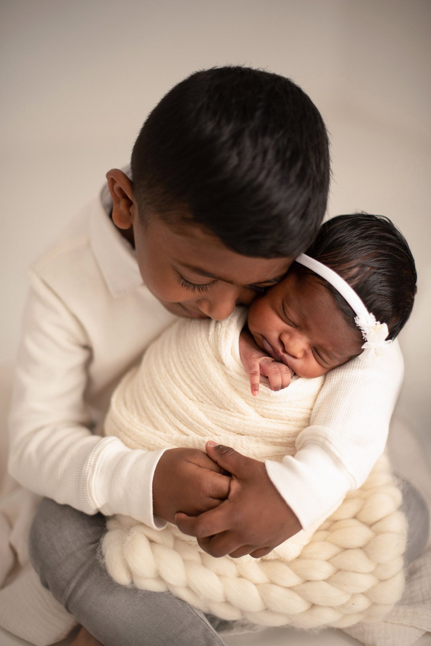 Young child holds sleeping newborn wearing headband nestled in cream knit blanket.