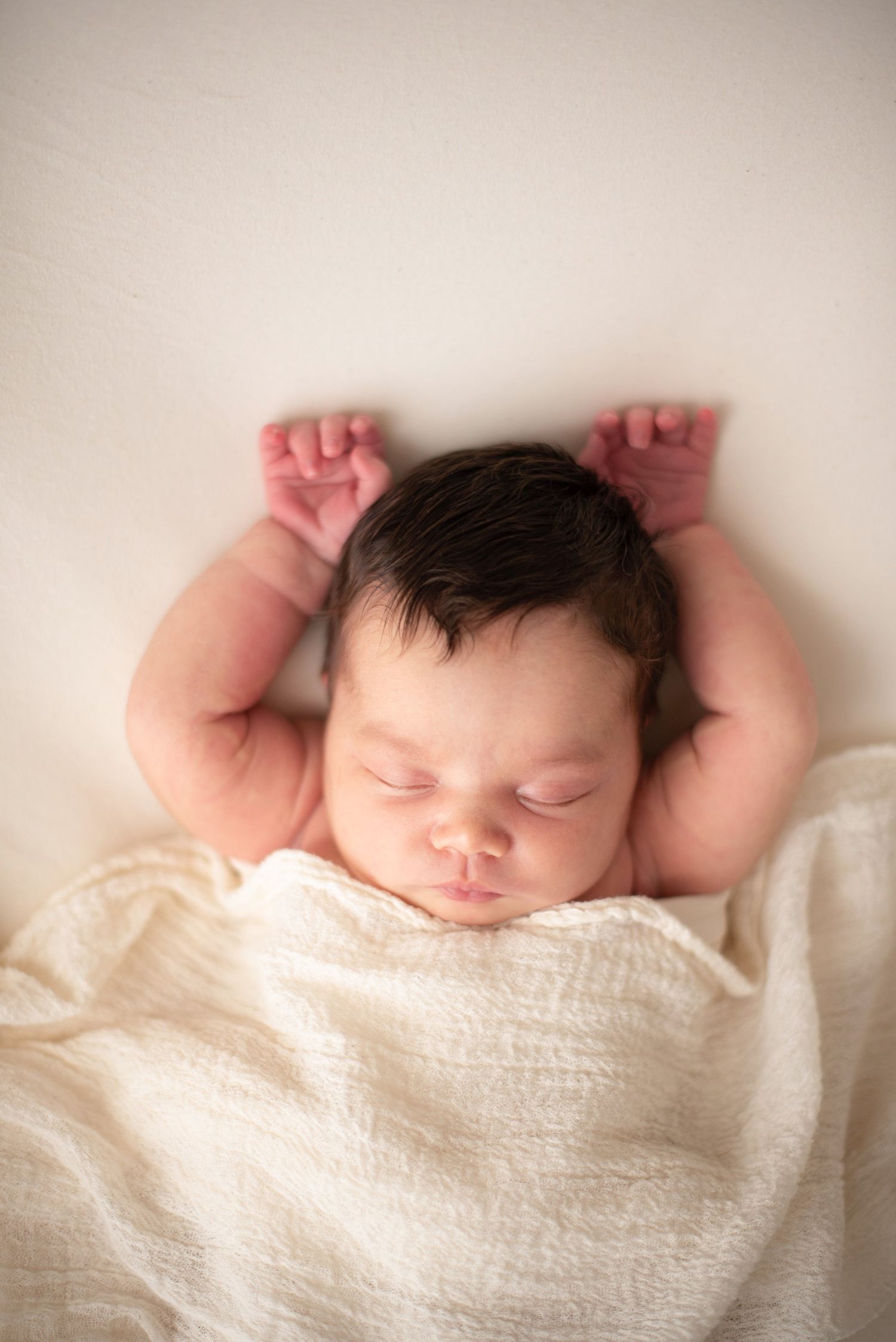 Sleeping newborn with arms raised on cream blanket during portrait session.