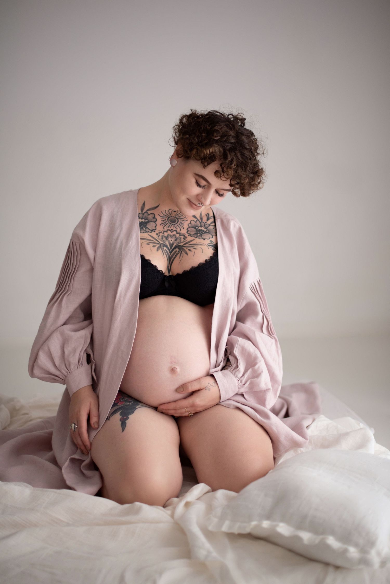 Artistic maternity photo showing pregnant person in pink robe and black lingerie against neutral backdrop.