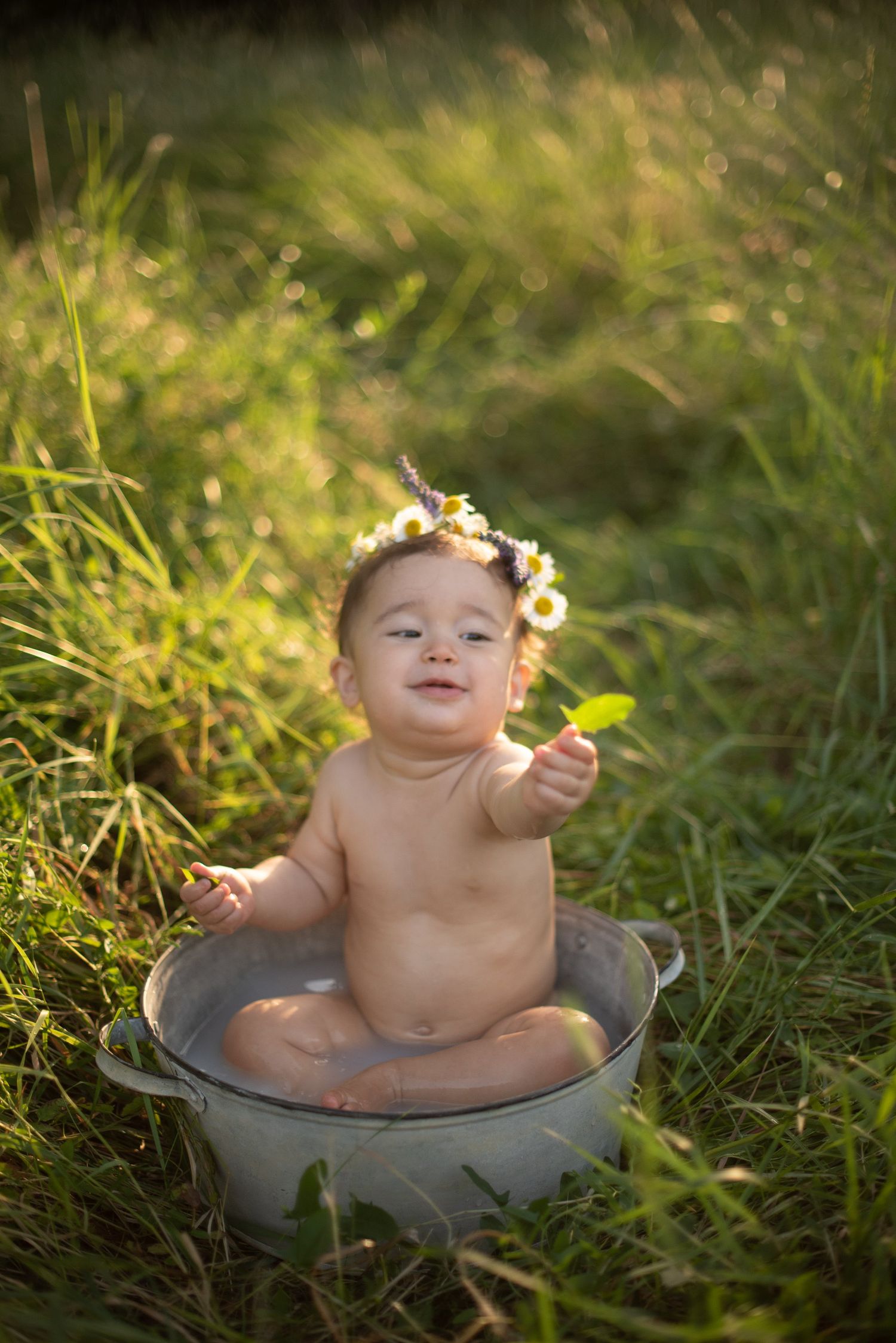 A baby wearing a floral crown sits in a metal basin outdoors surrounded by sunlit grass.