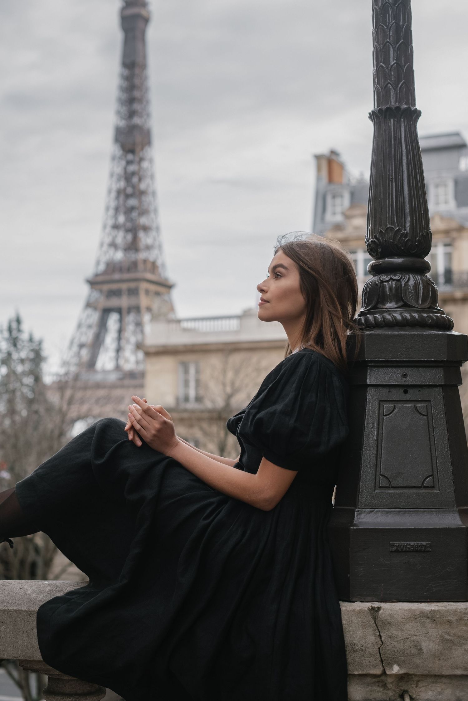 A woman in a black dress sits contemplatively against a lamppost with the Eiffel Tower in view.