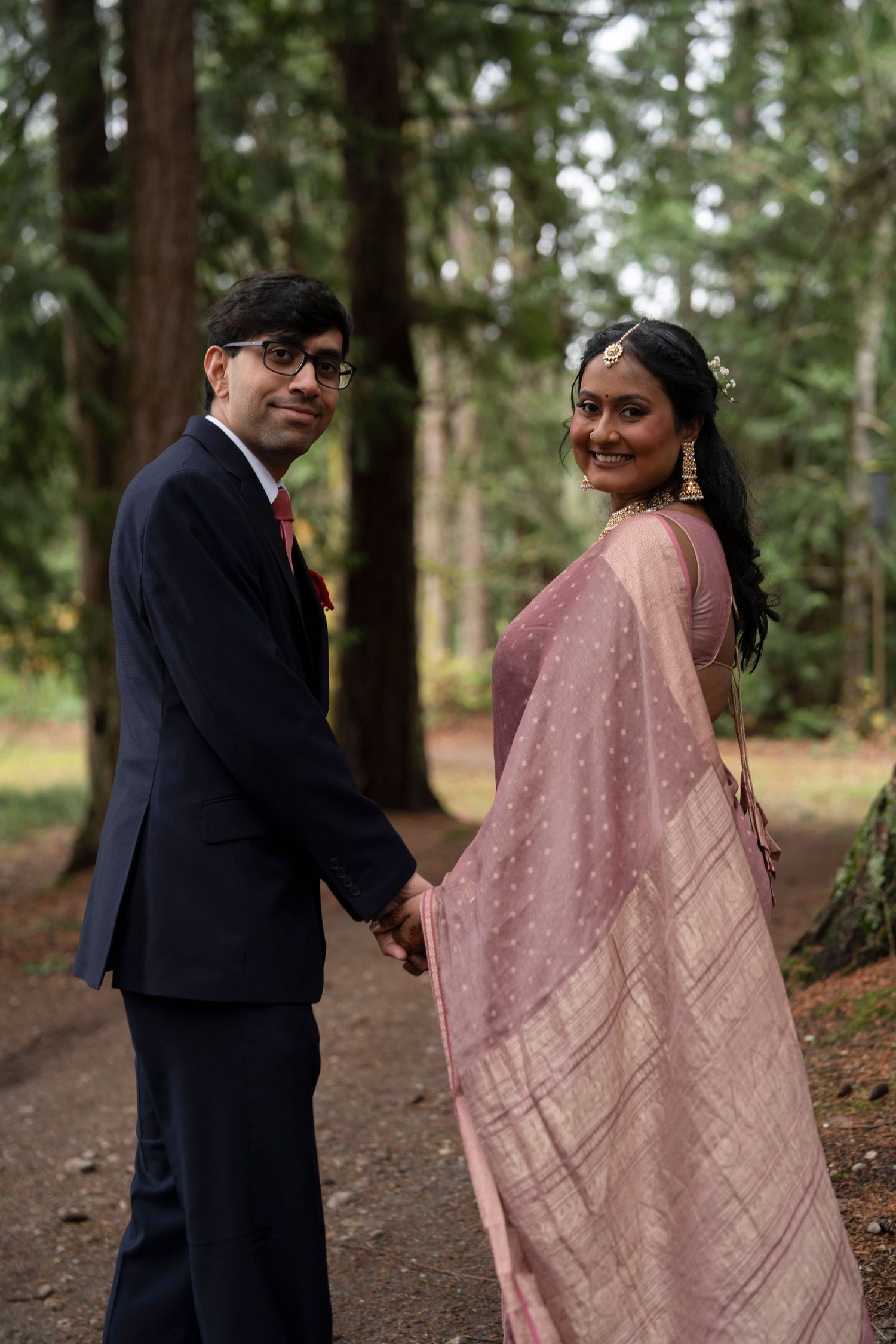 A couple in formal attire stands together on a forest path, with the person wearing a flowing pink sari.