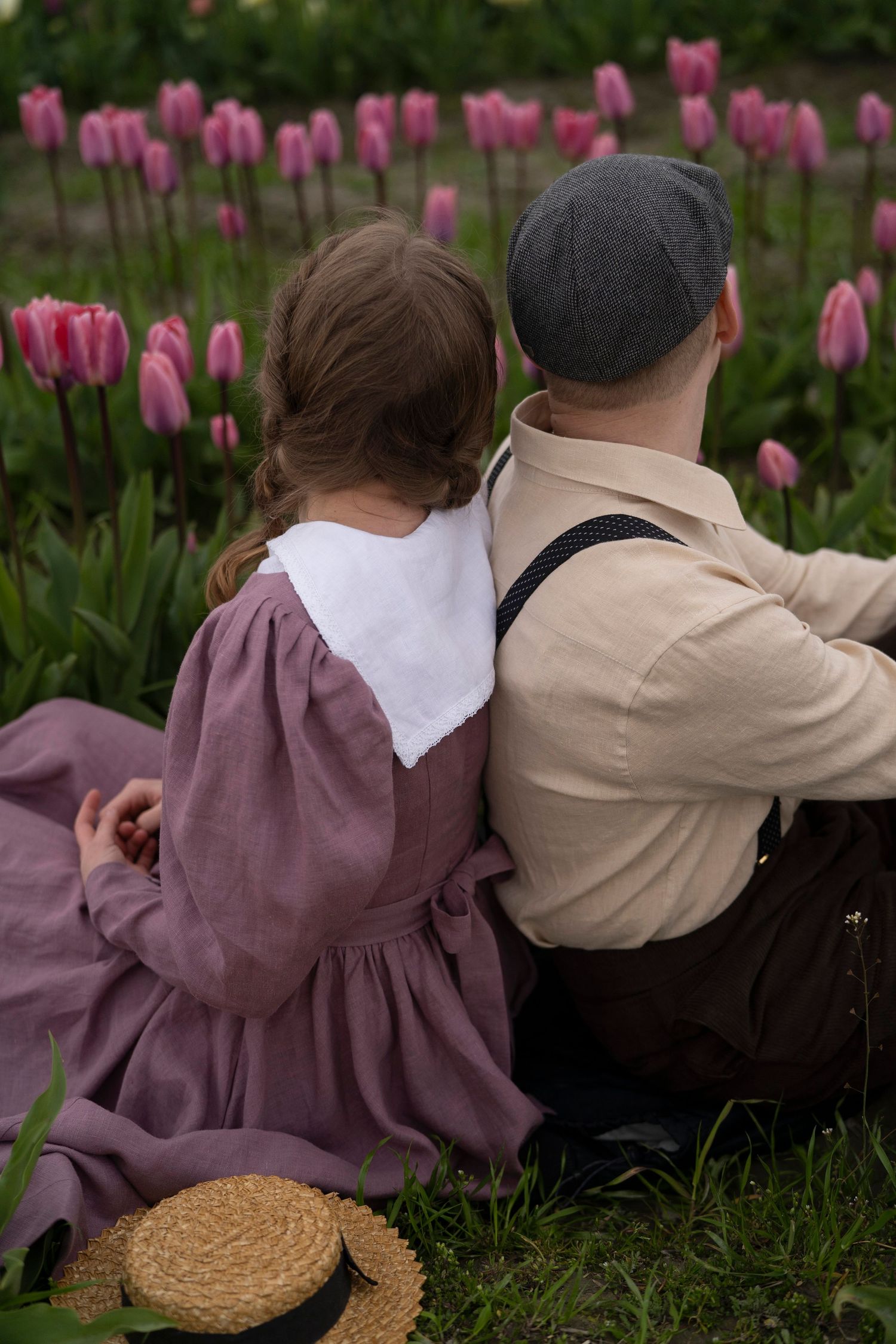 A couple sits together in vintage clothing near a garden of blooming pink tulips with a straw hat resting beside them.