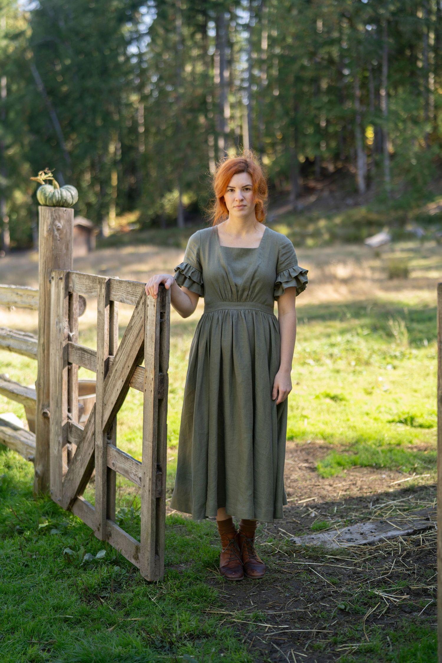 Woman in olive green prairie dress standing by rustic wooden gate.