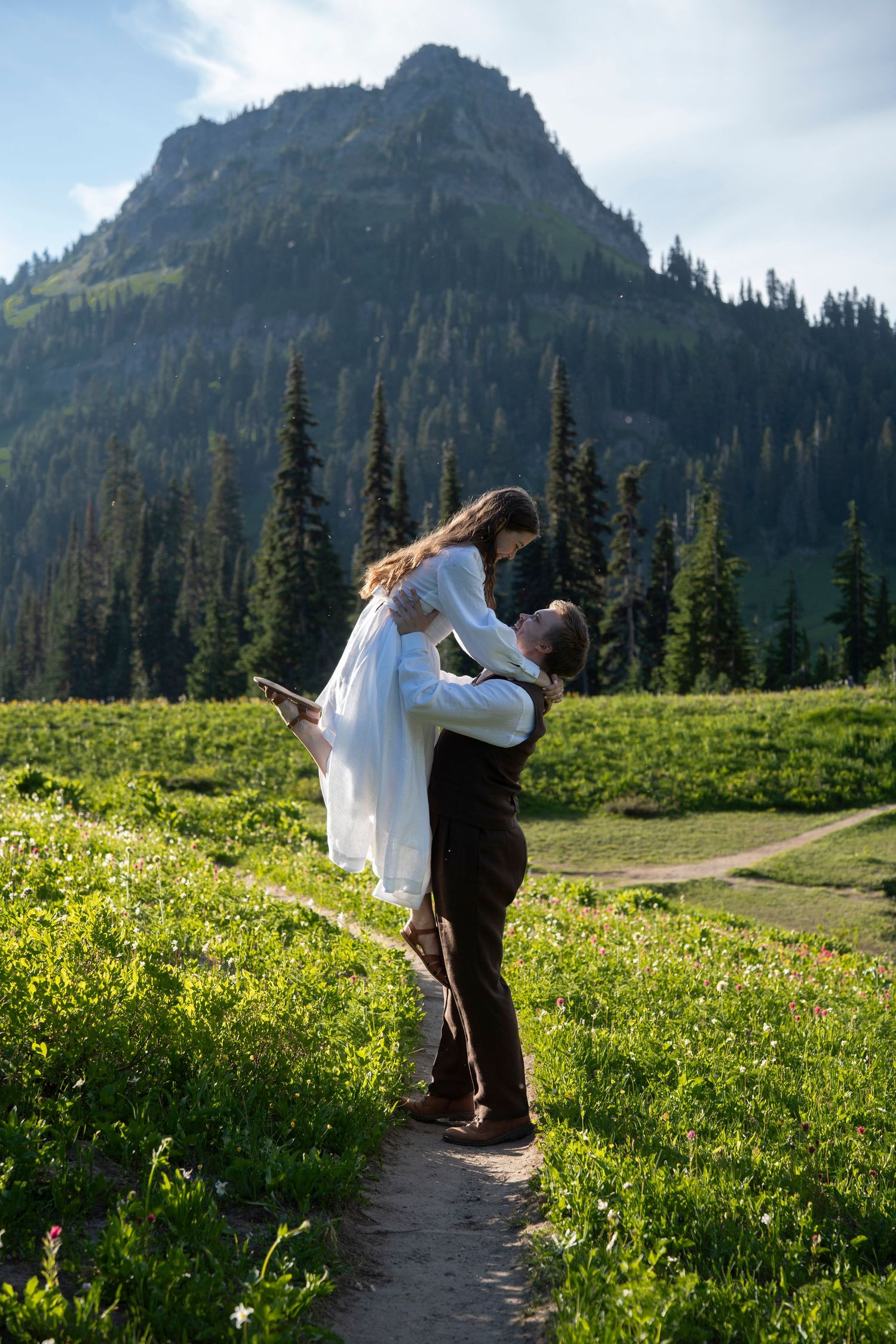 A couple embraces on a mountain trail surrounded by wildflowers and evergreens with a majestic peak in the background.