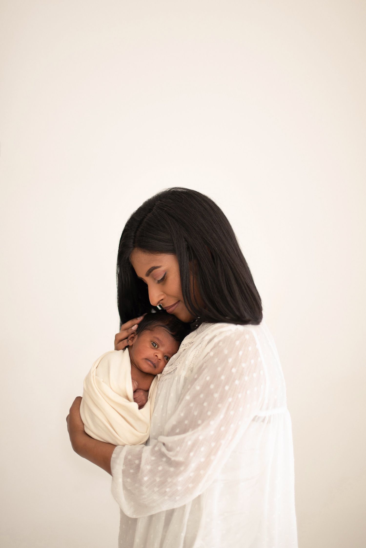 Parent in white sheer top embraces their sleeping newborn swaddled in cream fabric against light background.