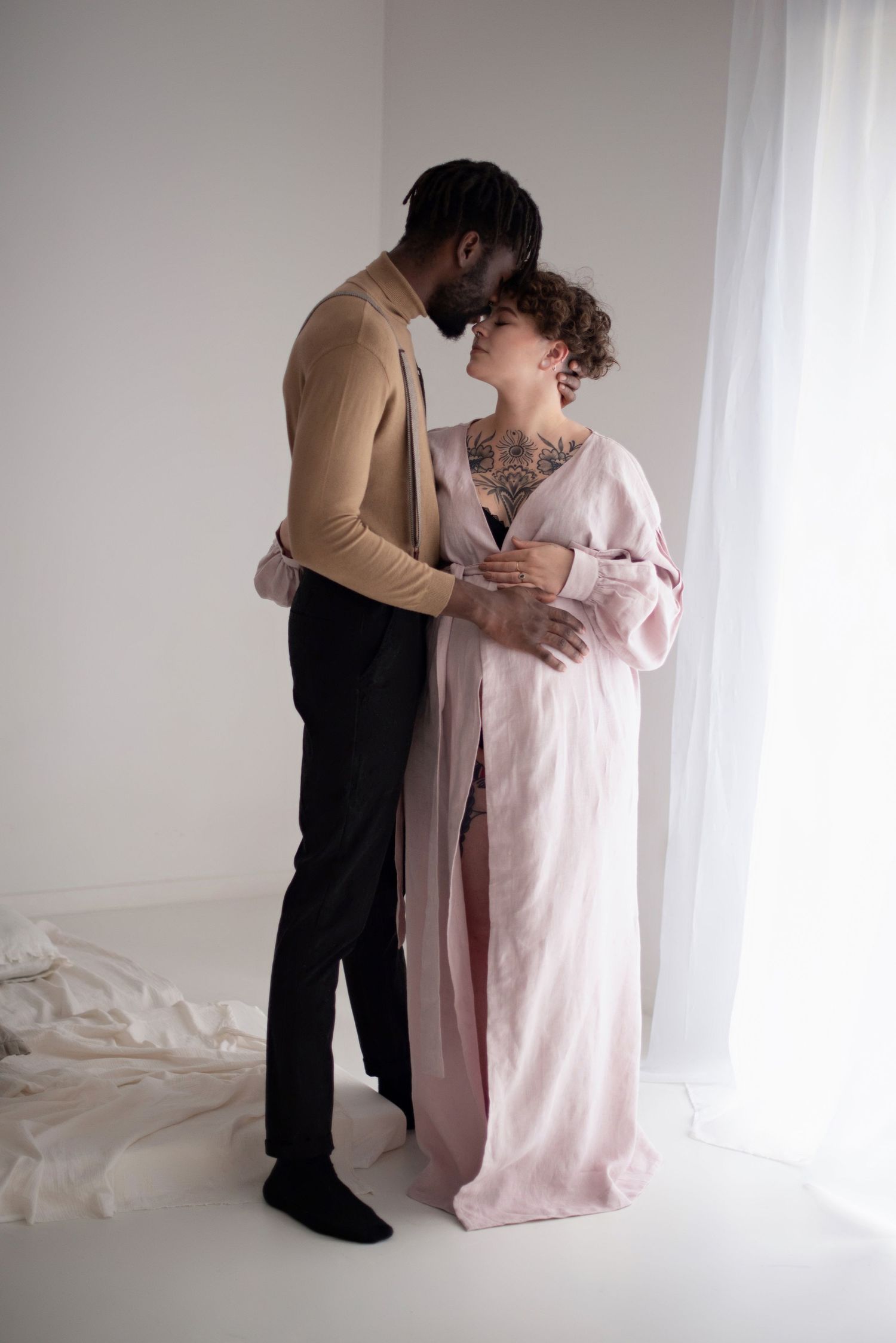 Tender moment between couple during maternity photoshoot with soft natural lighting by window.