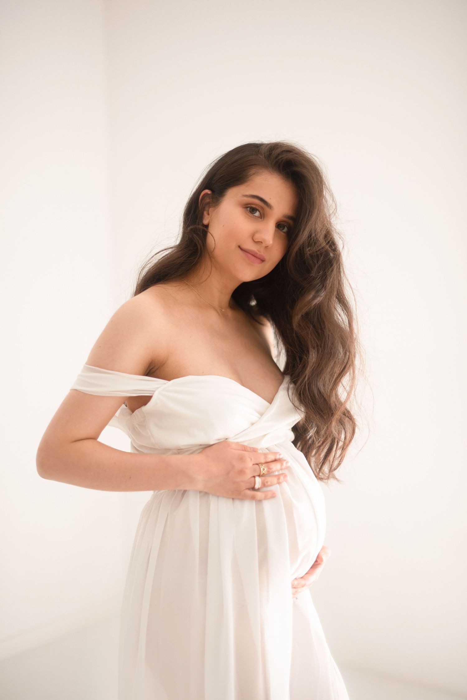 Elegant maternity portrait with woman in white off-shoulder dress against light background.