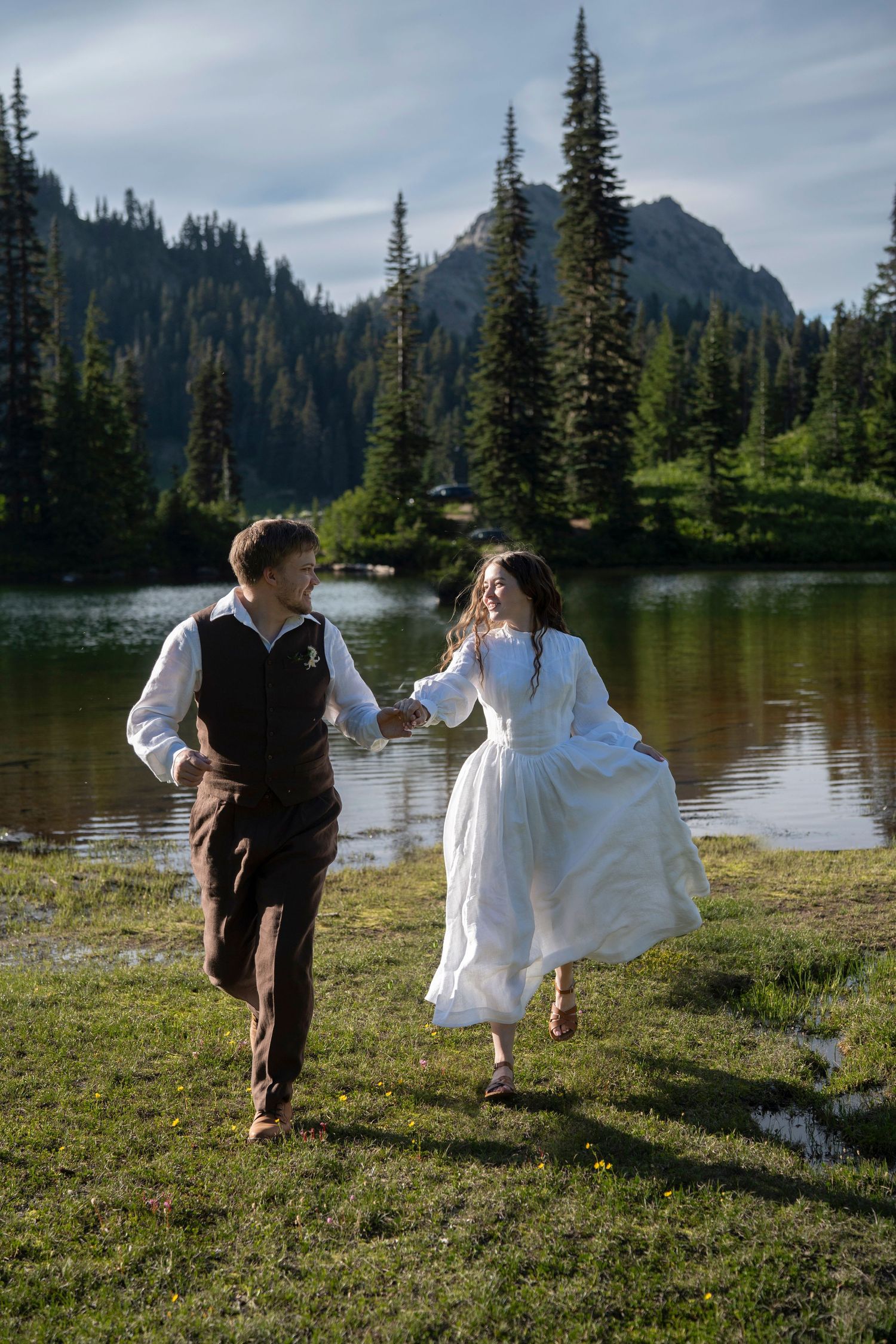 A couple runs together by an alpine lake surrounded by evergreen trees and mountains in the Pacific Northwest.