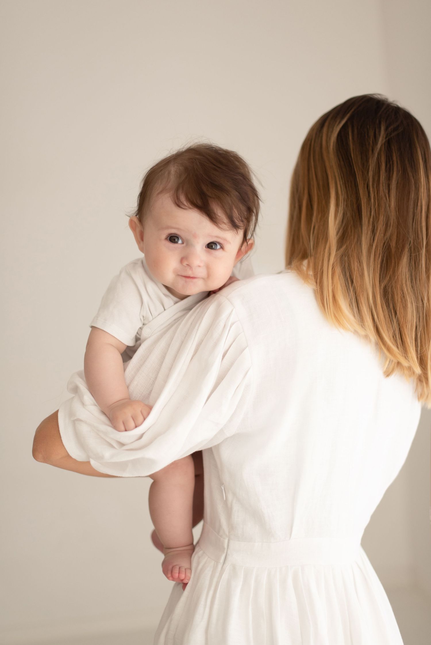 A baby in a white outfit peeks over the shoulder while being held by someone wearing a white dress.