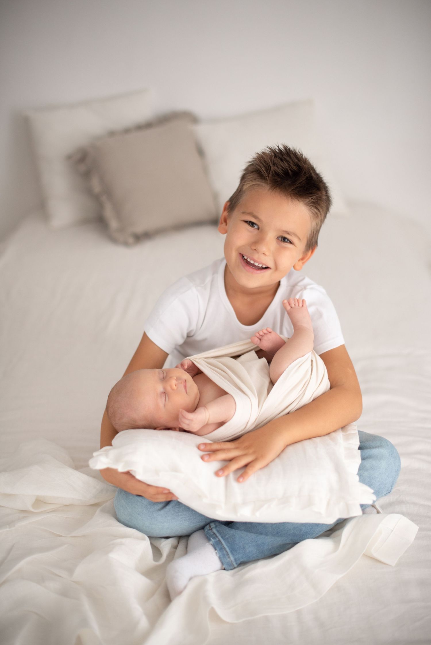 A young boy in white sits on a bed holding a newborn baby sibling.