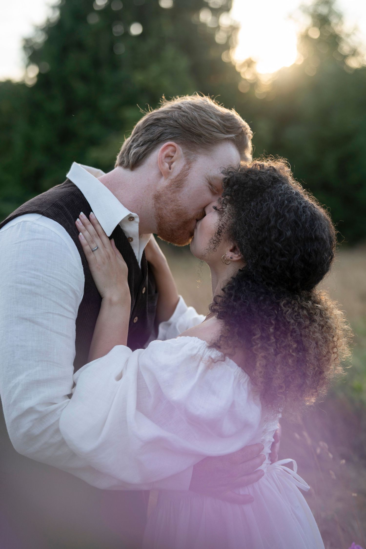 A romantic couple shares an intimate kiss outdoors at sunset with golden light filtering through the trees.