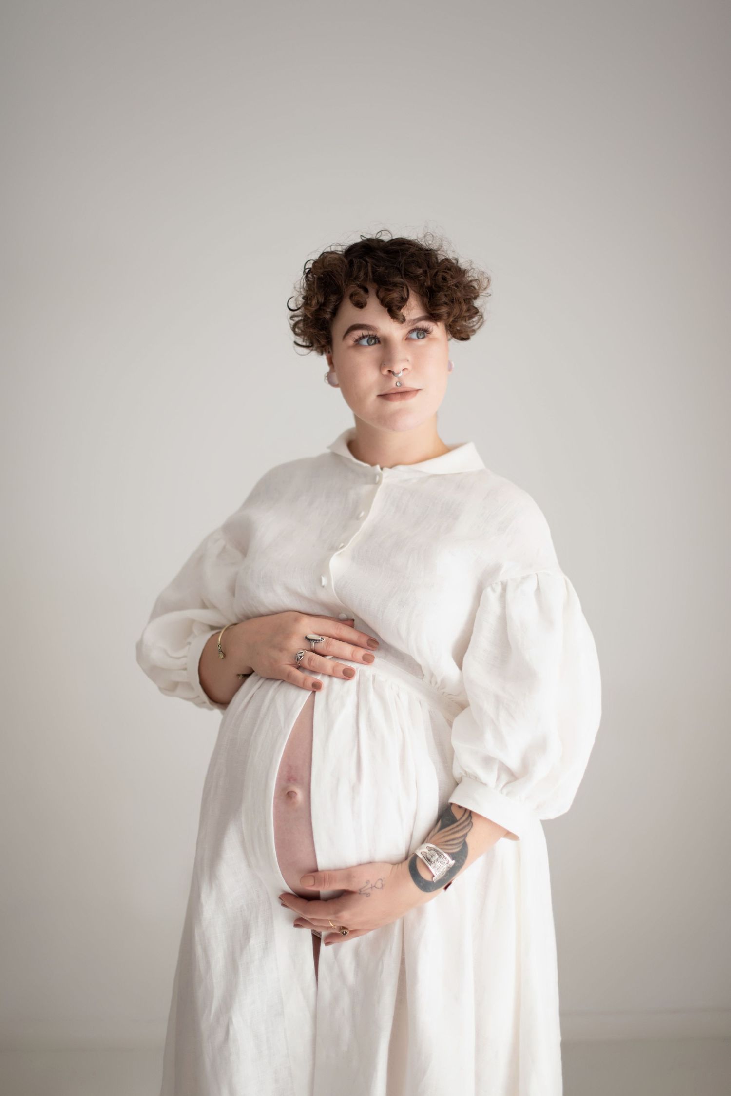 Person wearing white dress holds pregnant belly while posing against light backdrop.