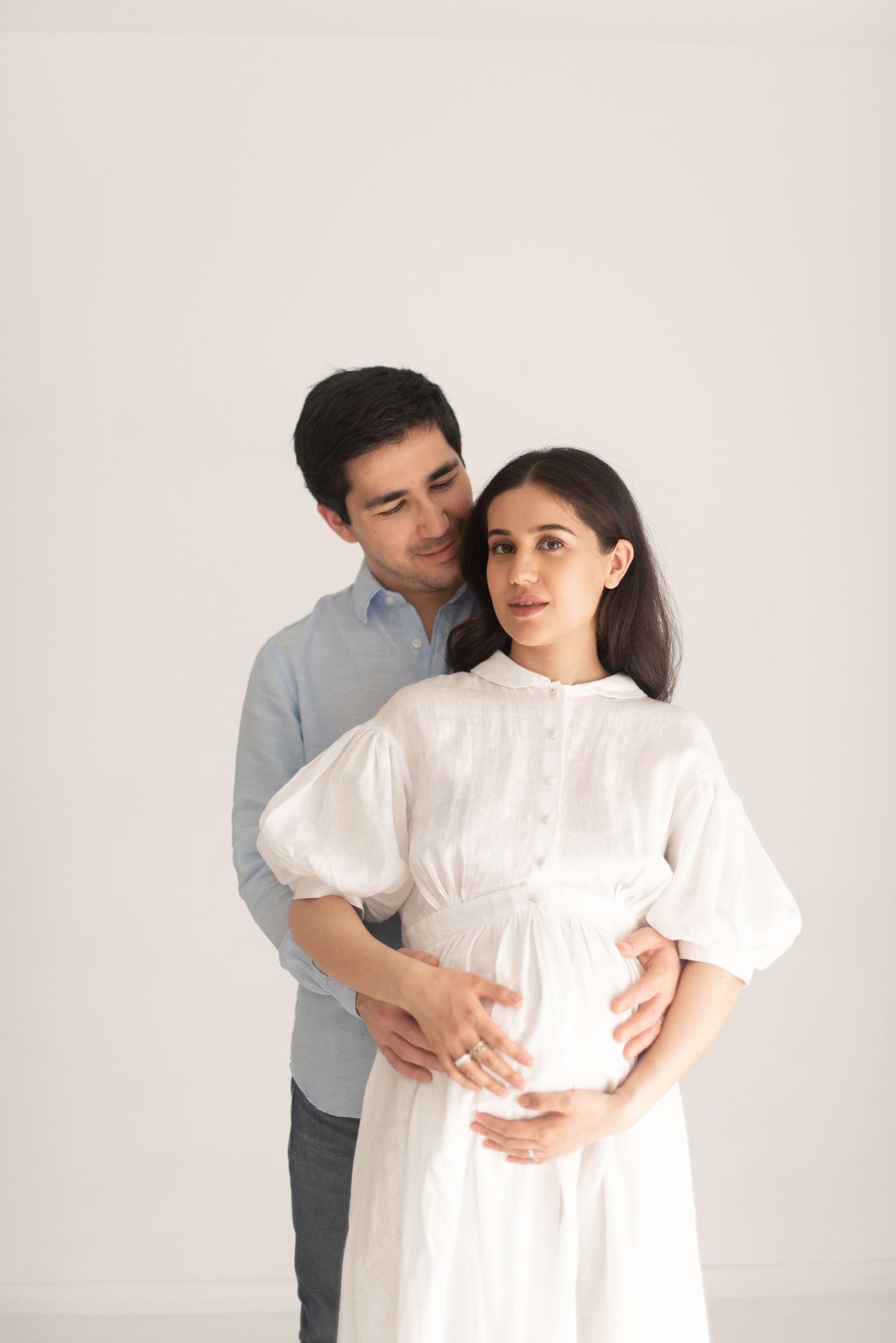 Romantic maternity photoshoot with couple embracing against white background, wearing white dress and blue shirt.
