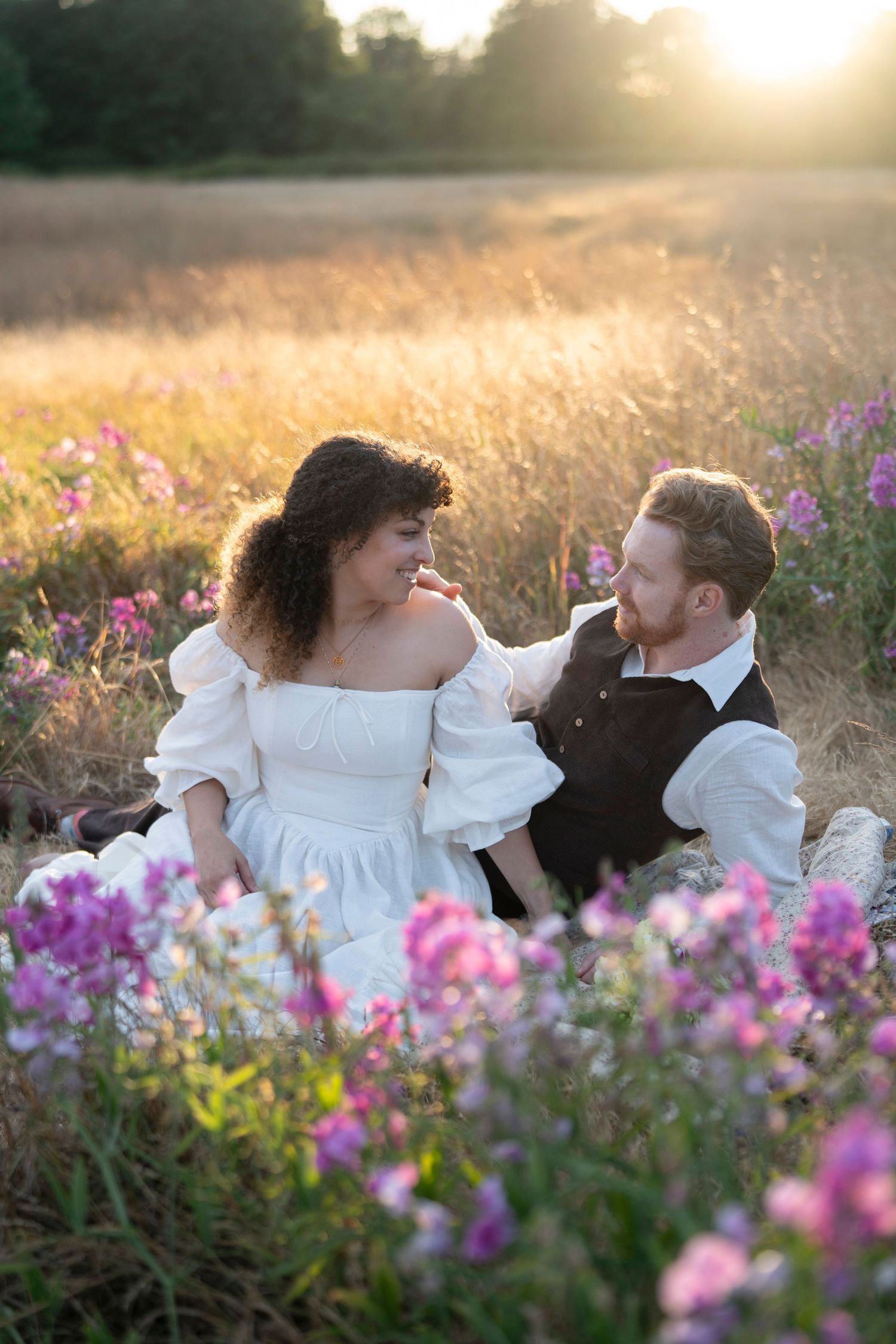 A couple embracing in a golden field at sunset with pink wildflowers in the foreground.