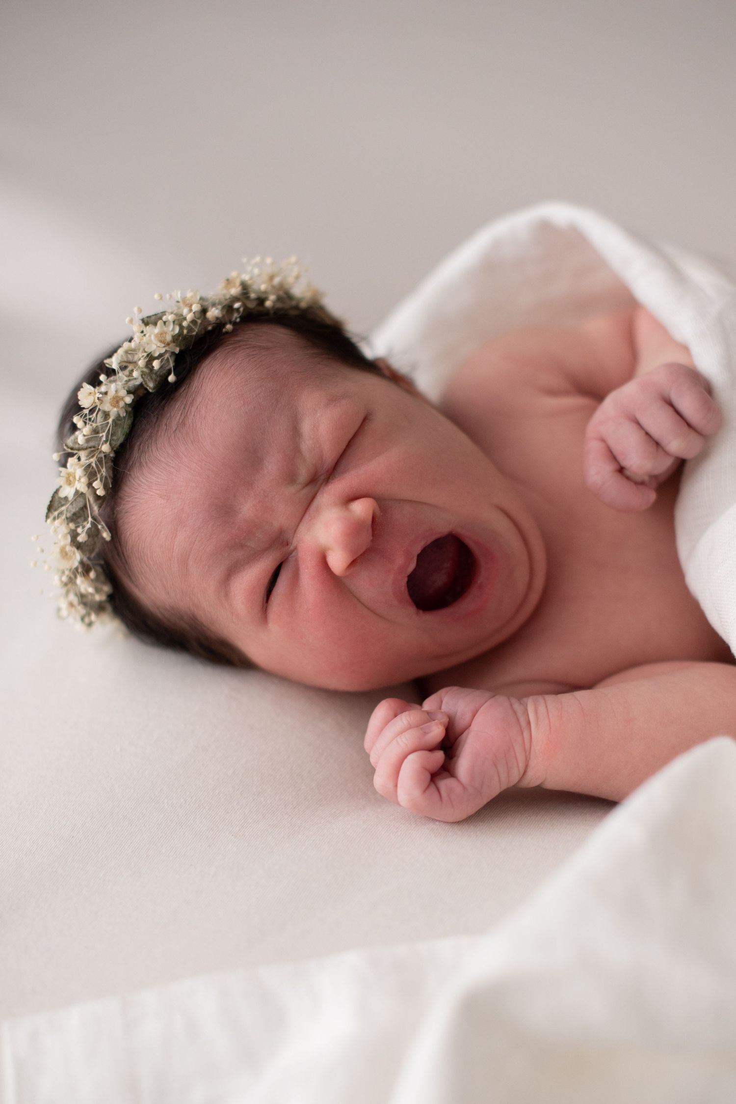 Newborn wearing floral crown yawning during professional photography session on beige backdrop.