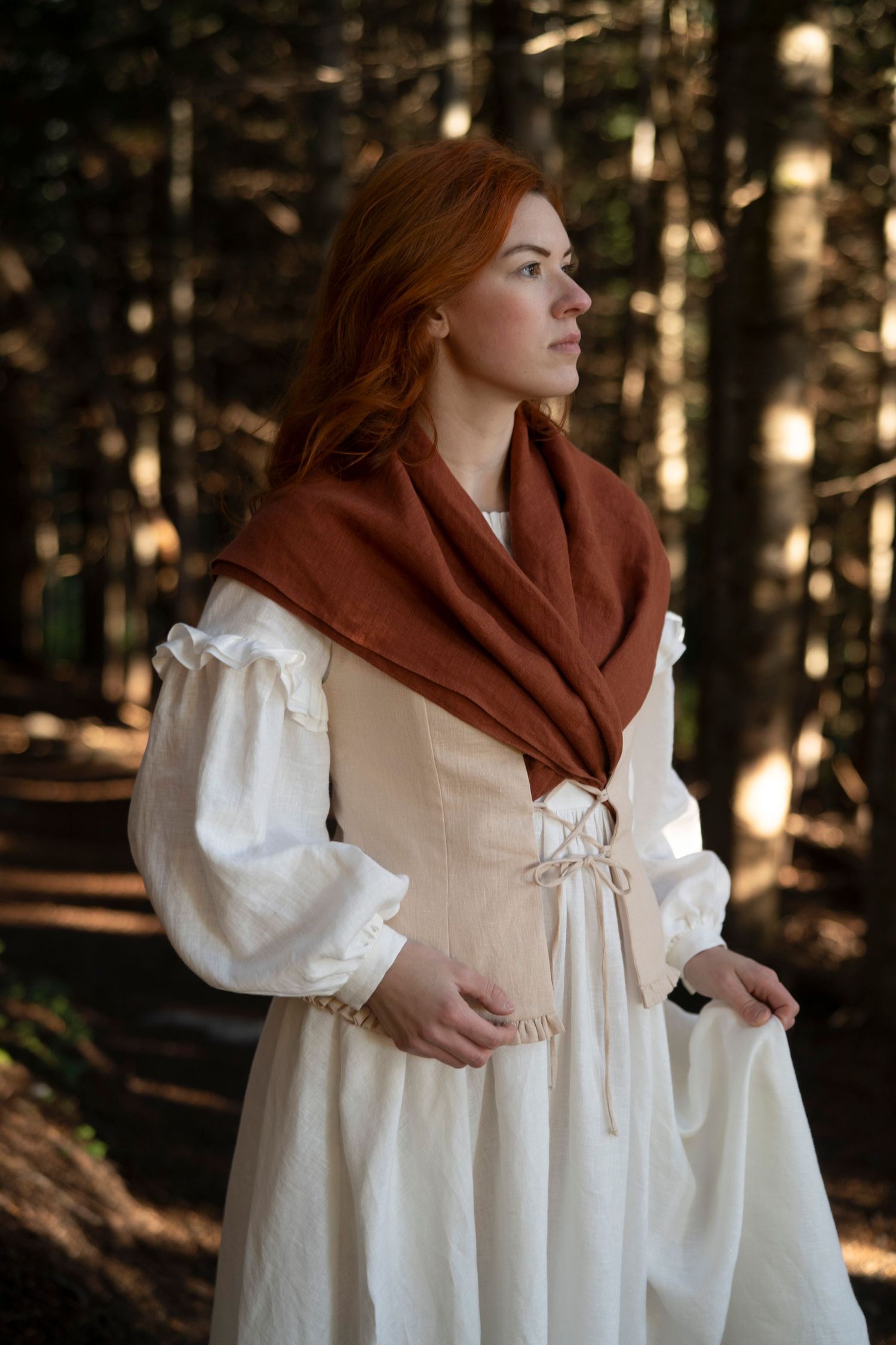 A figure in period attire wearing a cream dress and rust shawl stands contemplatively in a forest setting.