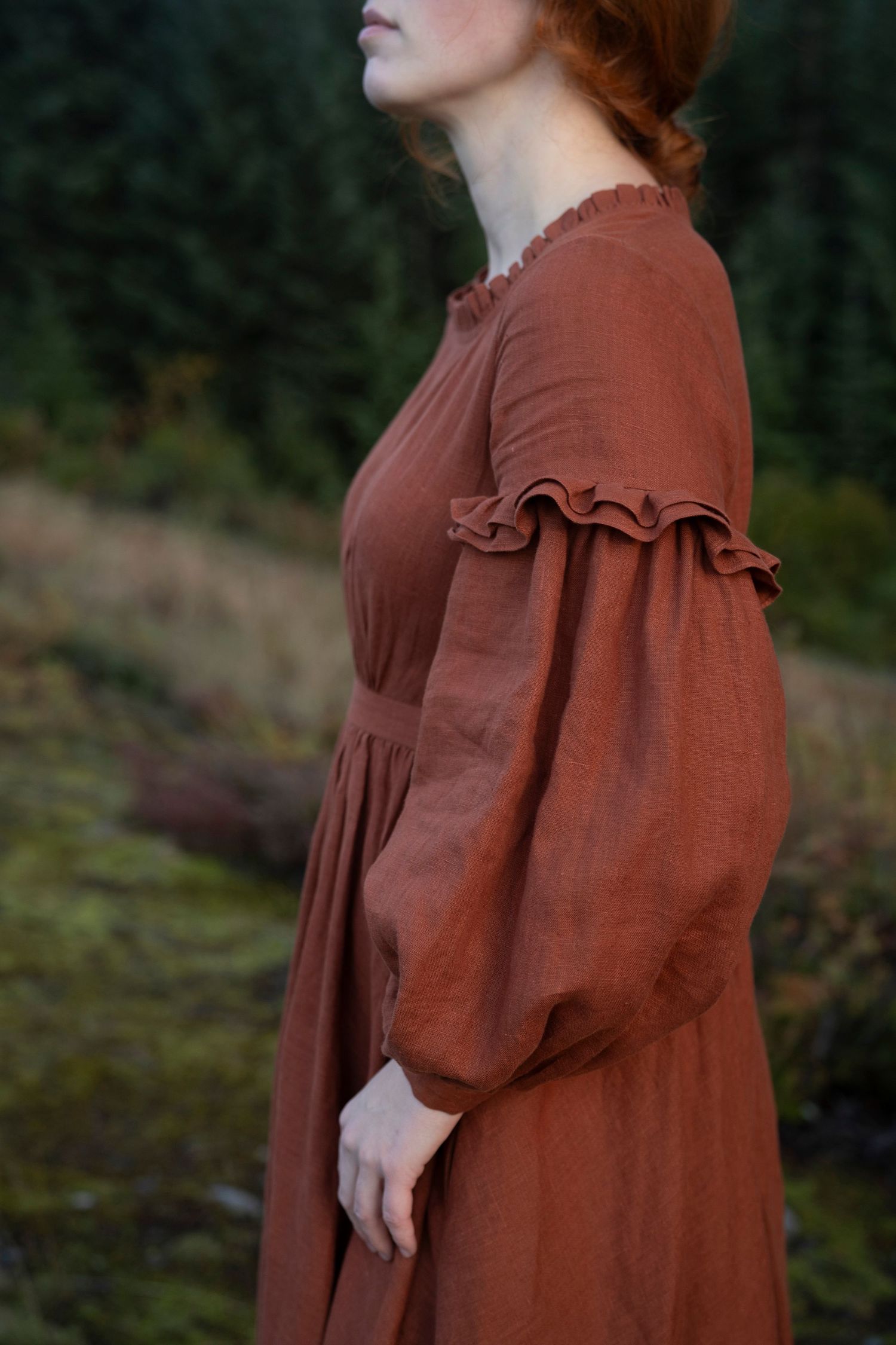 Close-up detail of a rust-colored prairie dress with ruffled details and puffed sleeves against a forested backdrop.
