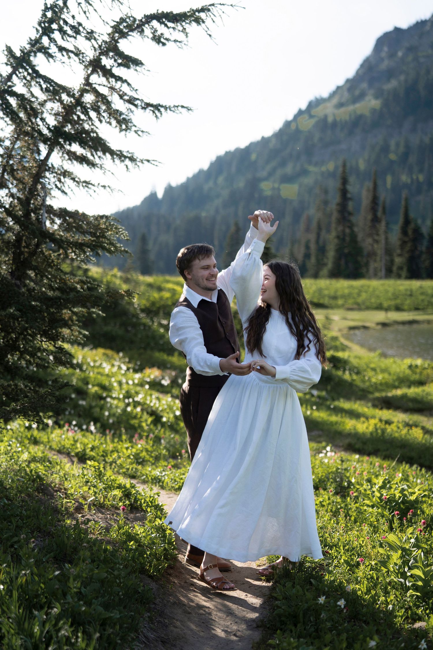 Two figures in white dresses dance together in a mountain meadow at sunset among wildflowers and evergreen trees.