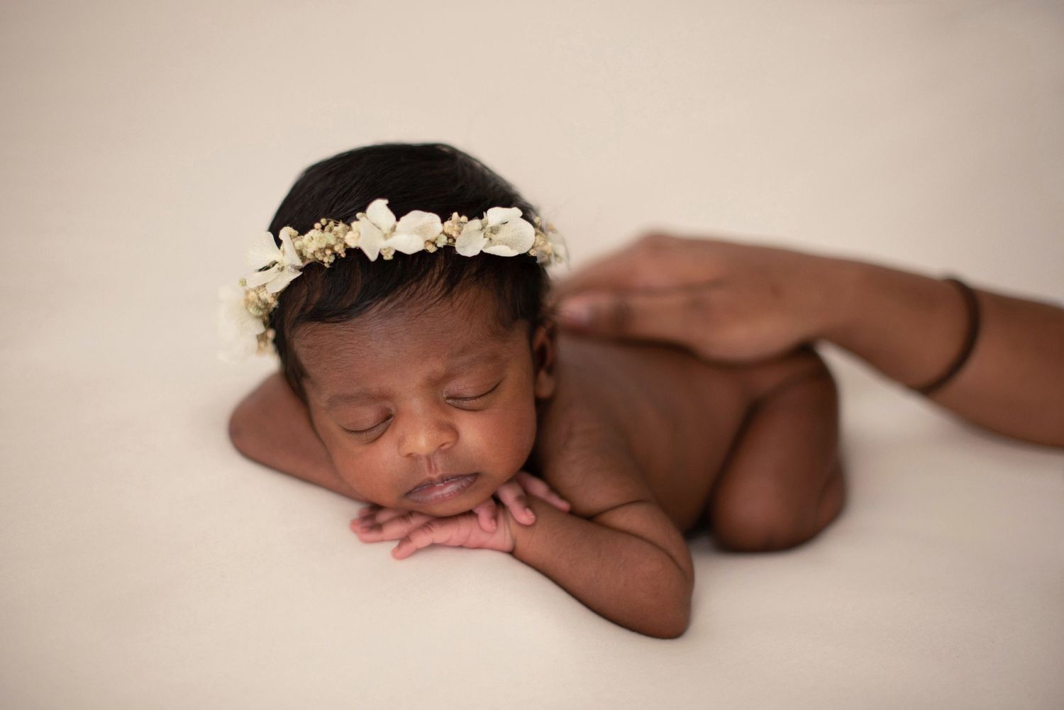 Sleeping newborn wearing floral crown poses on cream background during professional photoshoot.