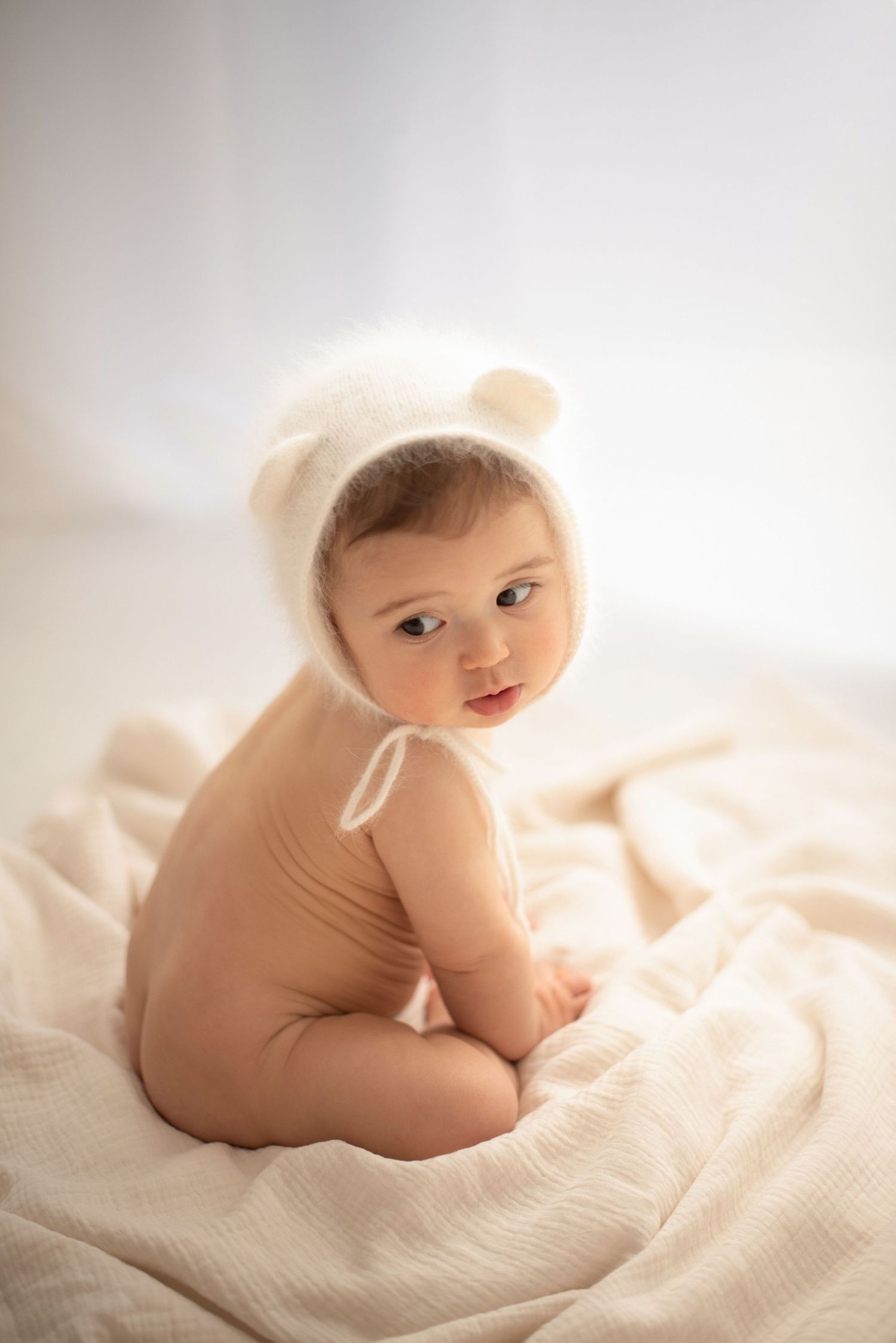 Baby wearing bear bonnet poses on cream blanket in gentle lighting.
