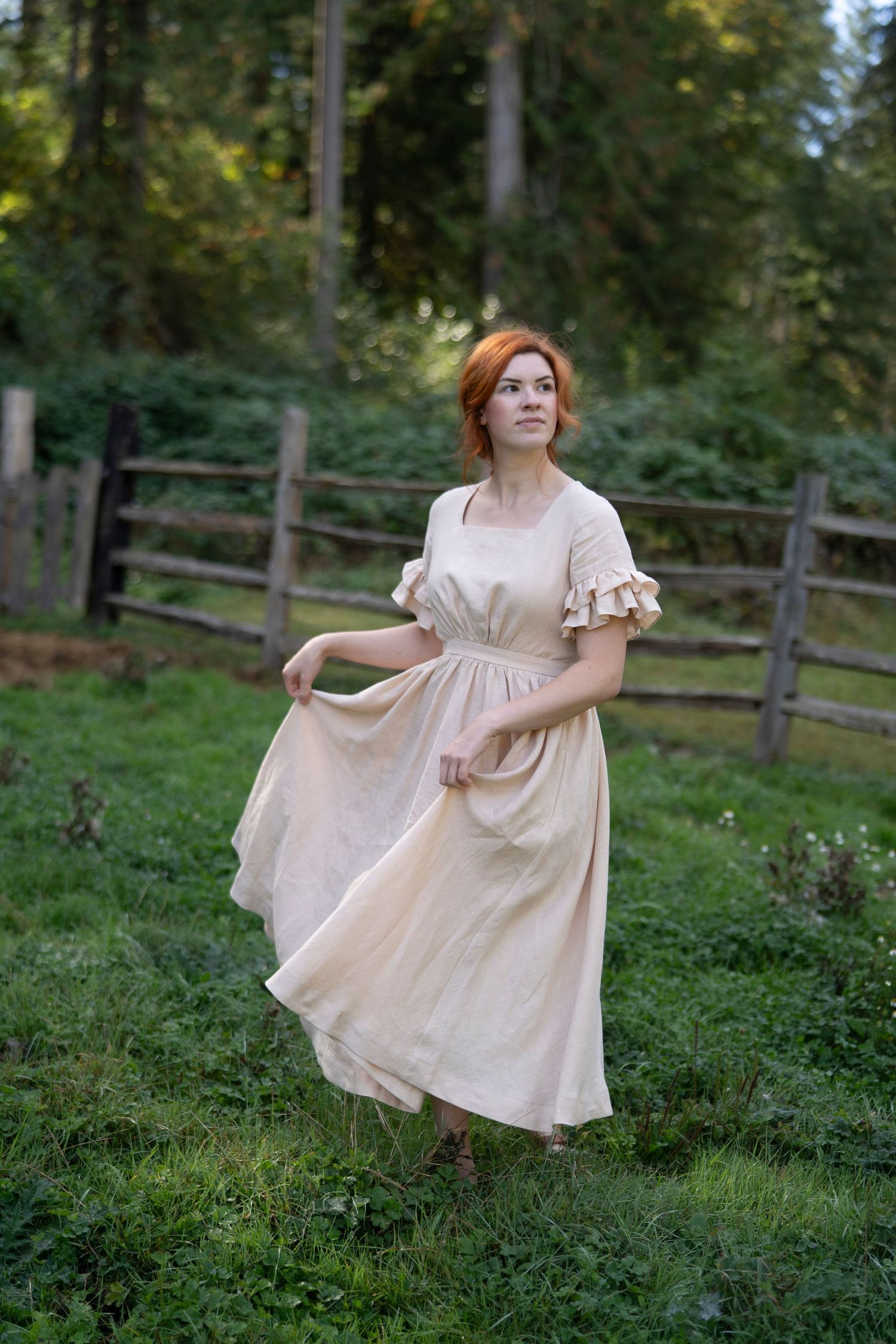A woman in a vintage beige dress twirls gracefully in a pastoral setting with wooden fences and trees in the background.