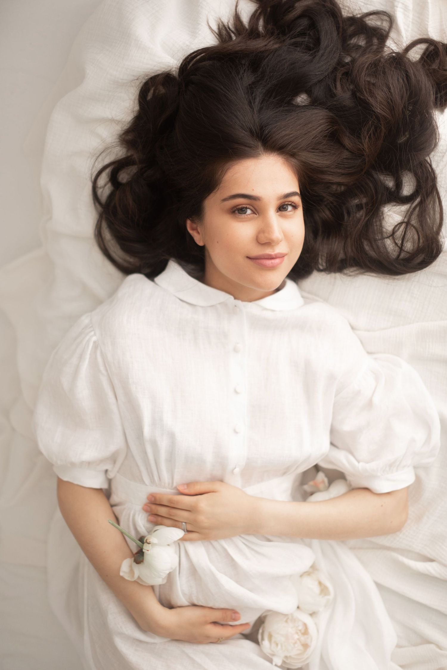 Woman in white dress with flowing dark hair lies on white fabric in artistic portrait.