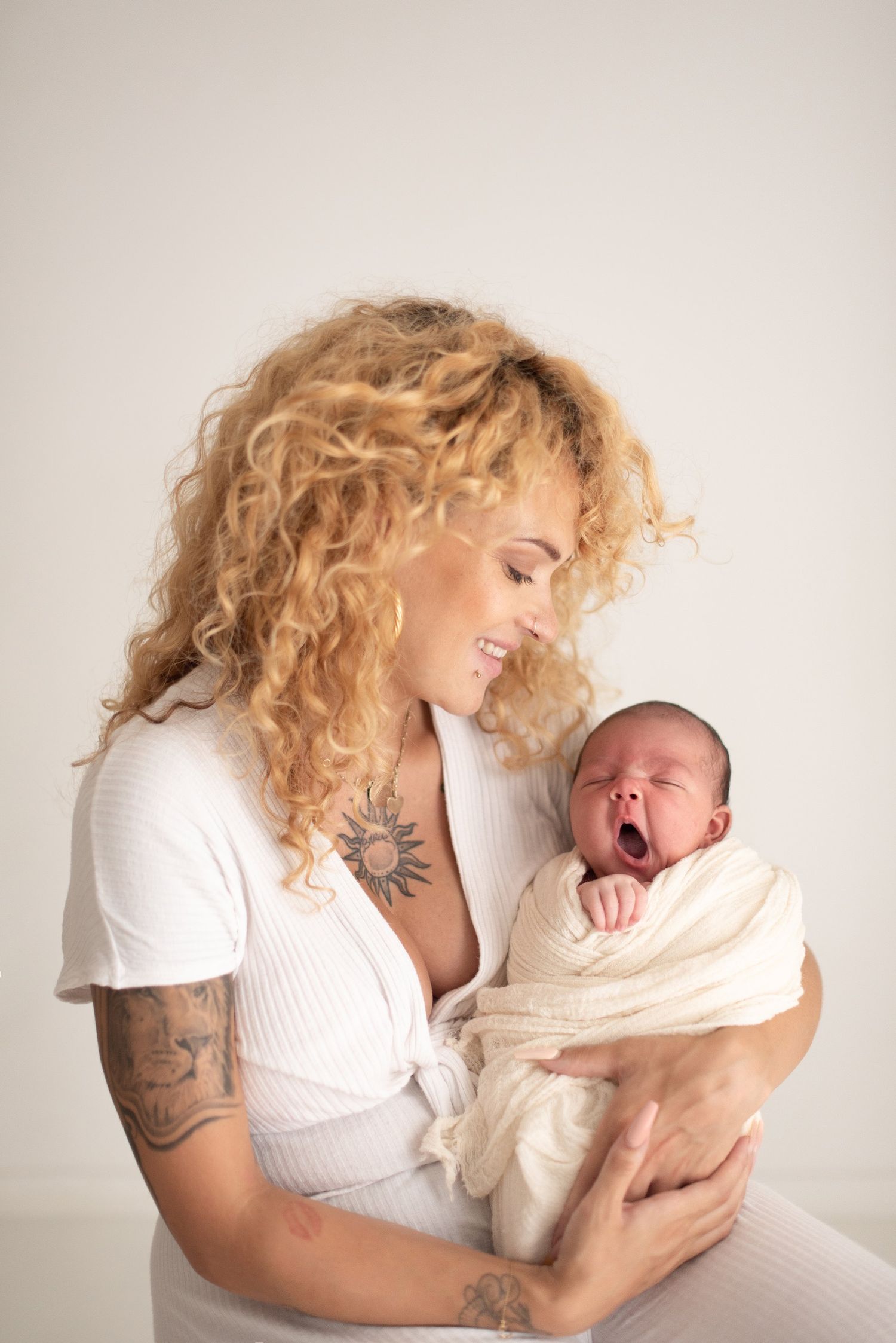 Parent with curly blonde hair and tattoos cradles a yawning newborn wrapped in white.