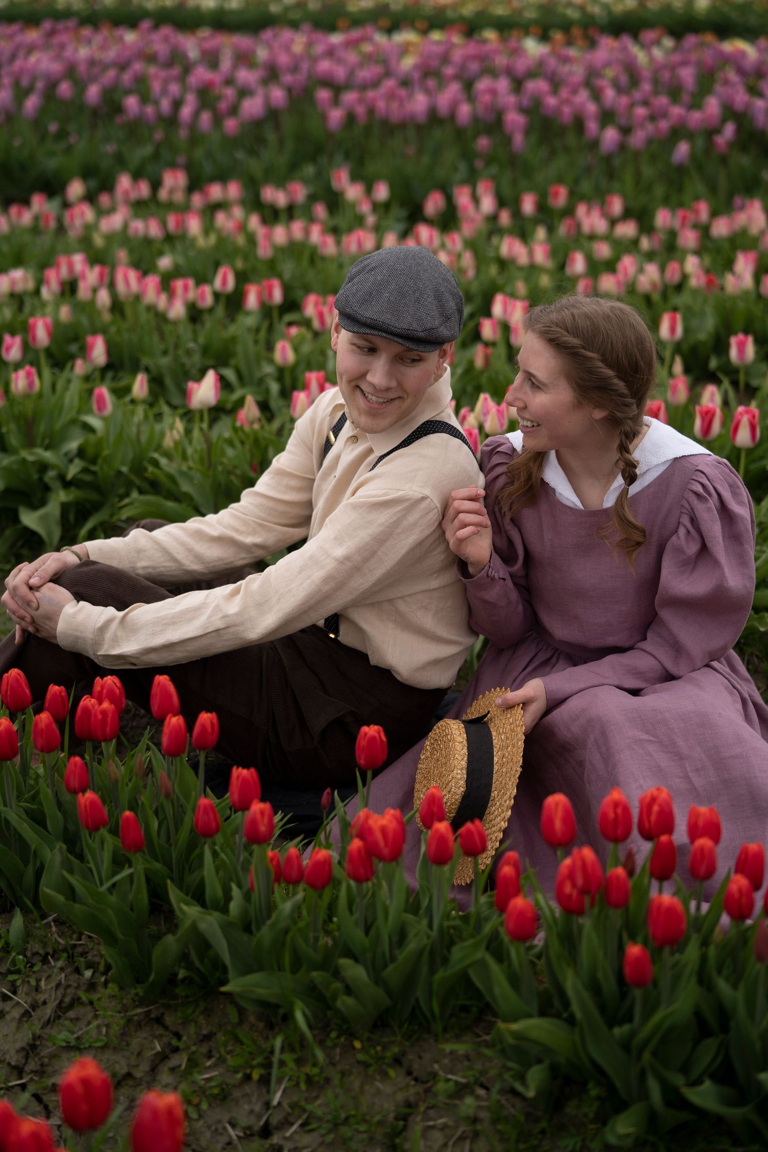 Two people share a romantic moment among vibrant pink and red tulip fields in vintage-style clothing.
