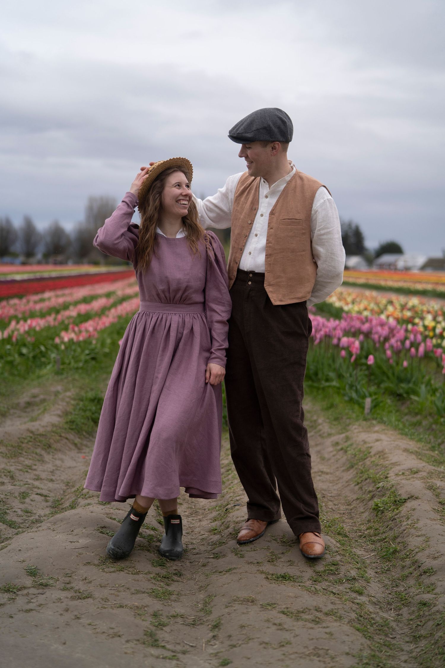 Two people in vintage clothing share a tender moment while walking along a path through colorful tulip fields at dusk.