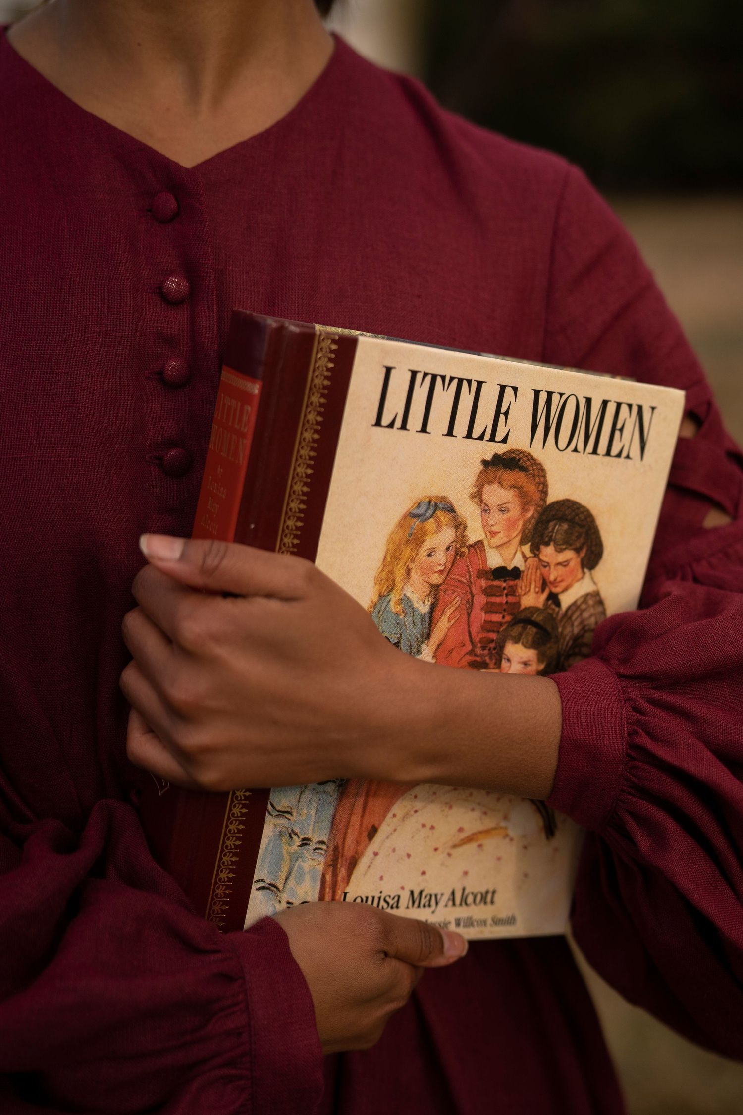 Close-up shot of hands holding a copy of 'Little Women' book while wearing a burgundy dress with buttons.