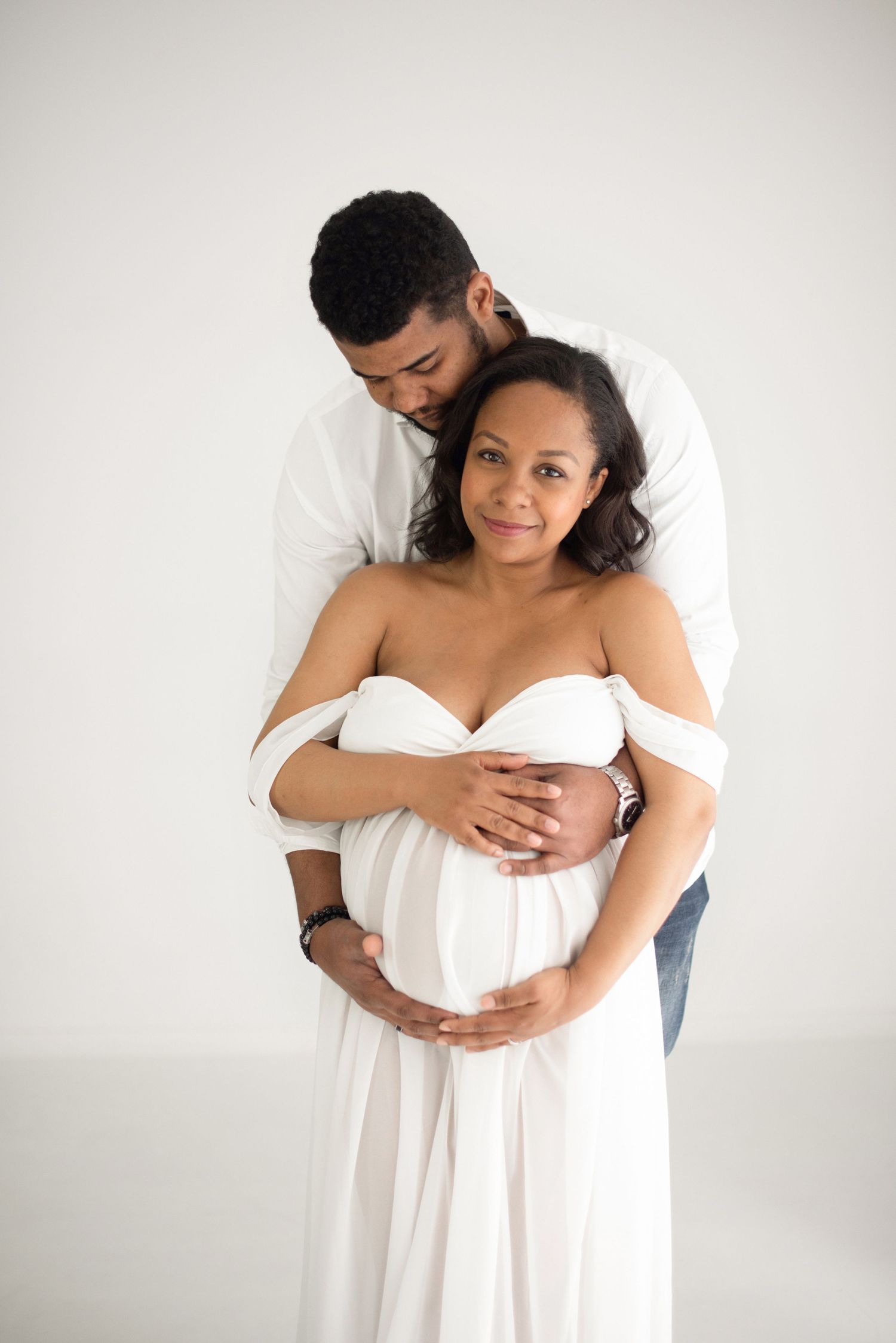 An intimate maternity portrait of a couple in white against a light background embracing tenderly.