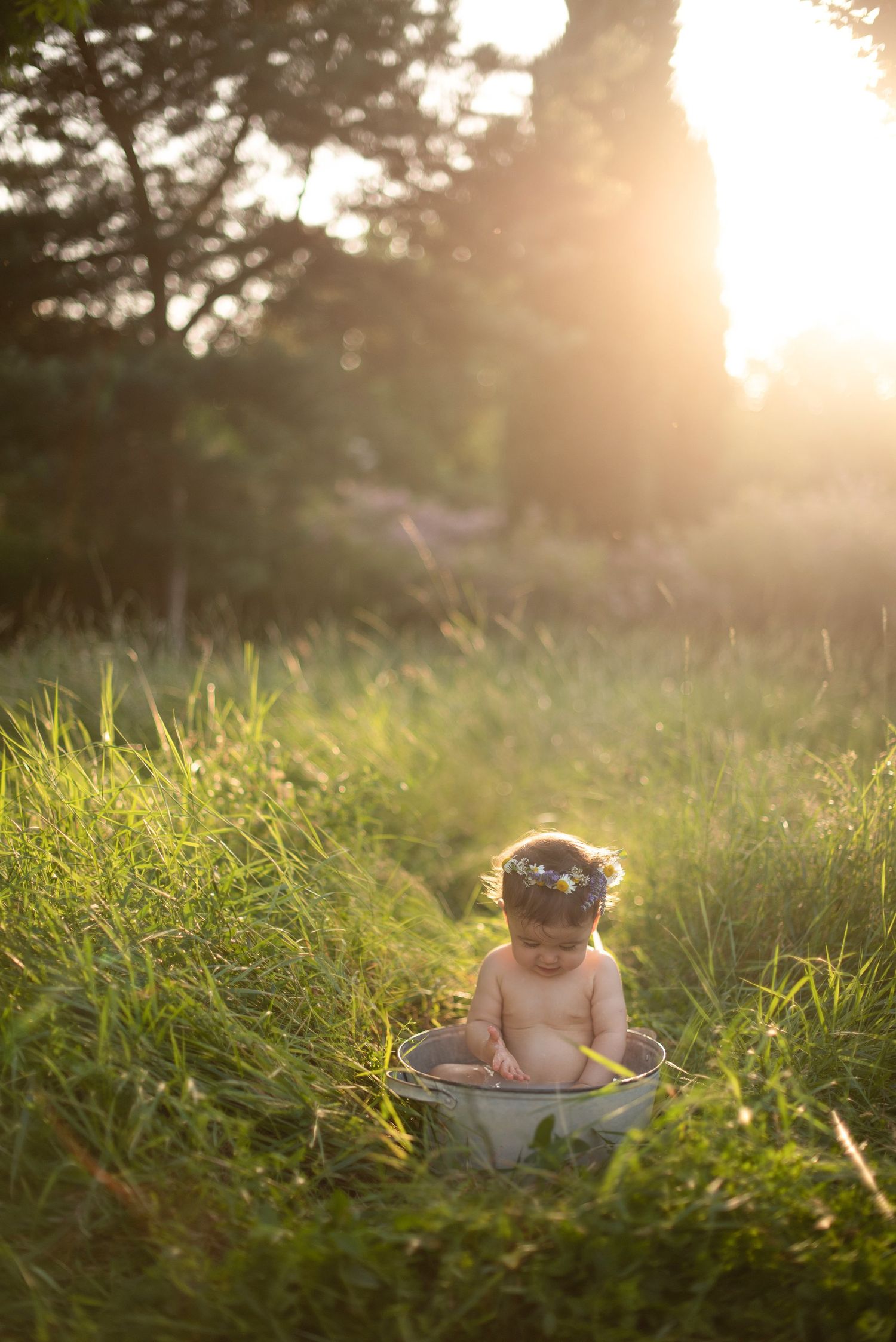 A baby bathes in a vintage metal tub amid tall grass during golden hour.