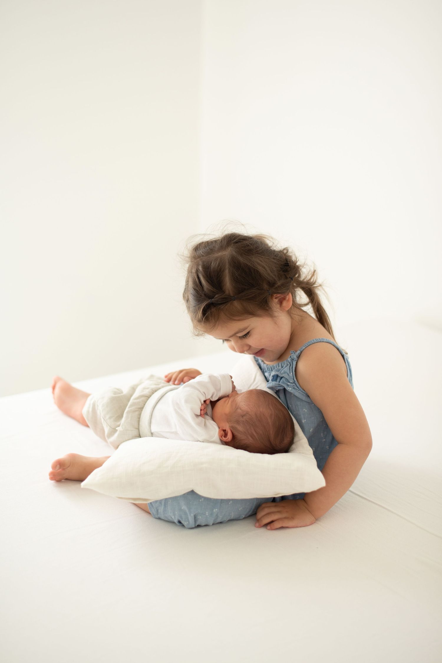 Young child in blue top tenderly watching over sleeping newborn on white pillow.