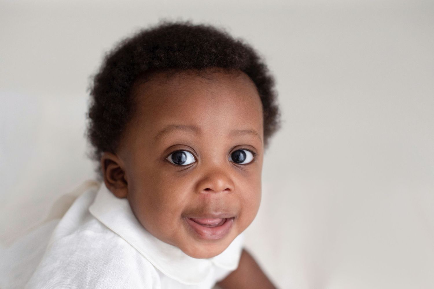 Adorable toddler wearing a white turtleneck smiles brightly against a neutral background.