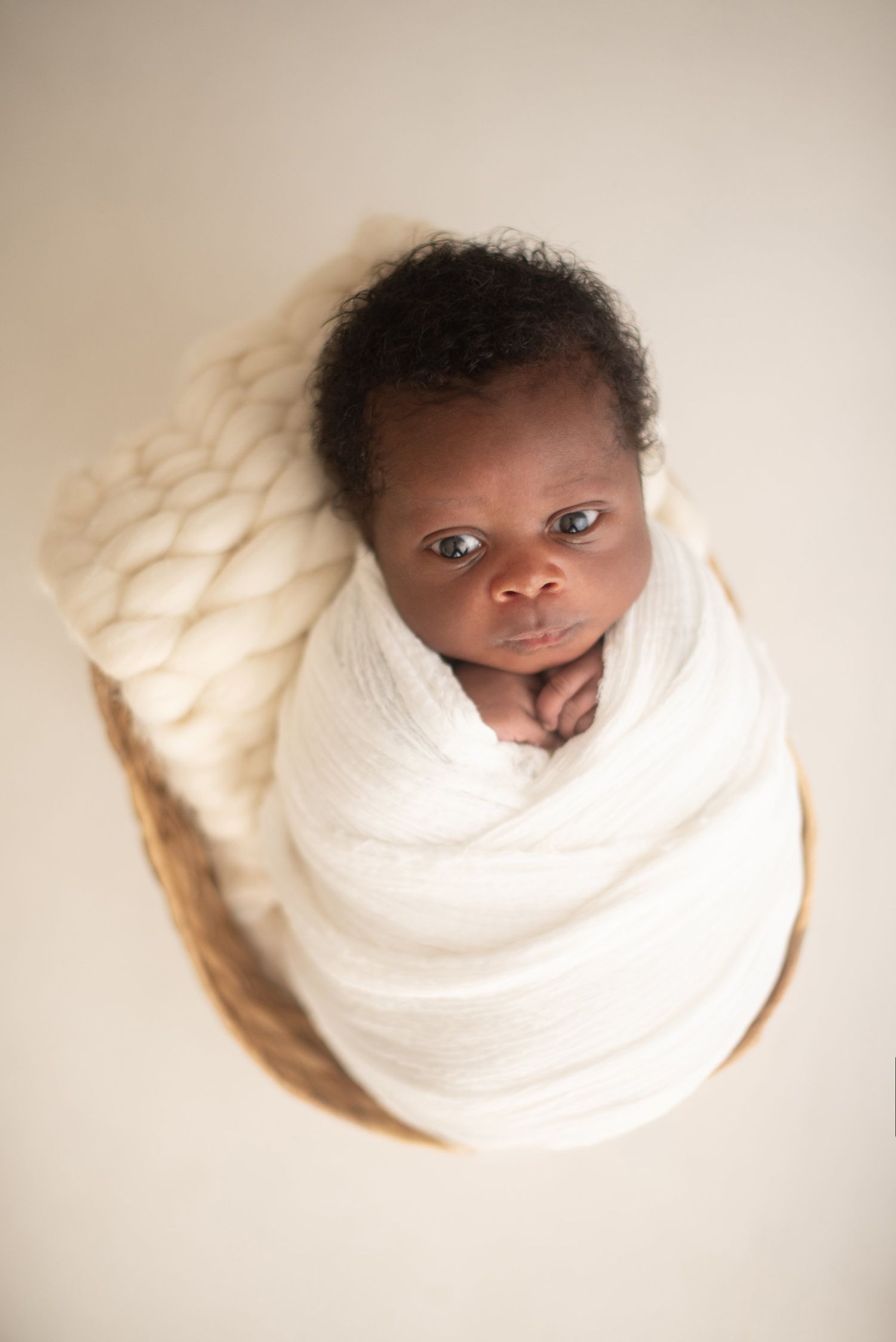 Overhead view of newborn baby wrapped in white blanket looking up at camera.