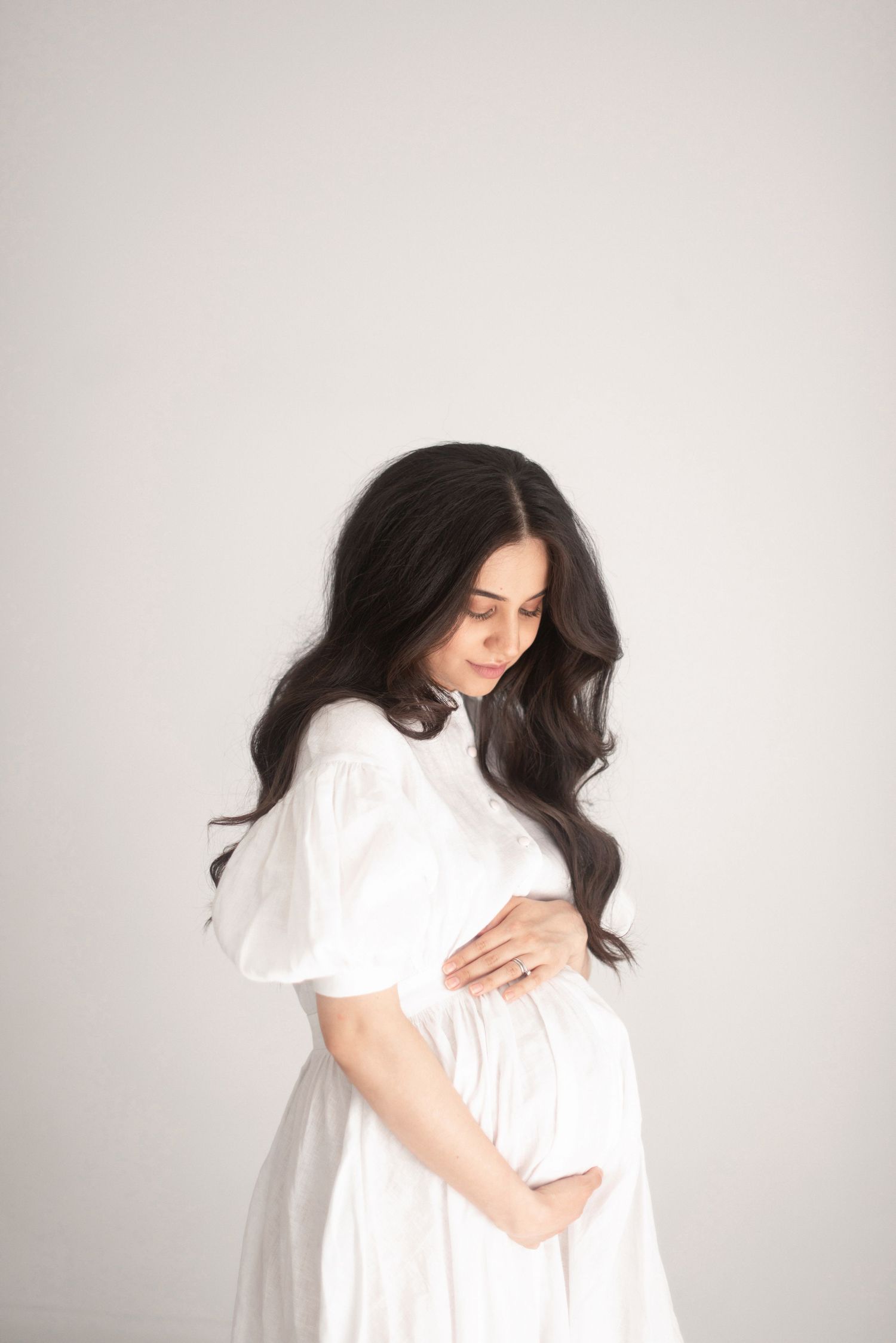 Dreamy maternity photo with long-haired woman in white dress looking down at baby bump.