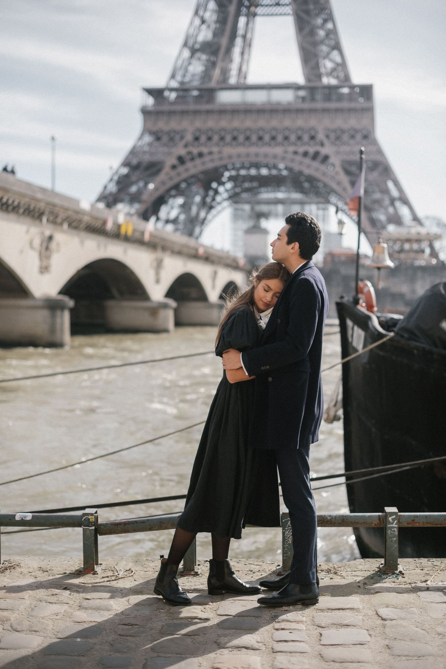 A couple embraces in front of the iconic Eiffel Tower on a cloudy day.