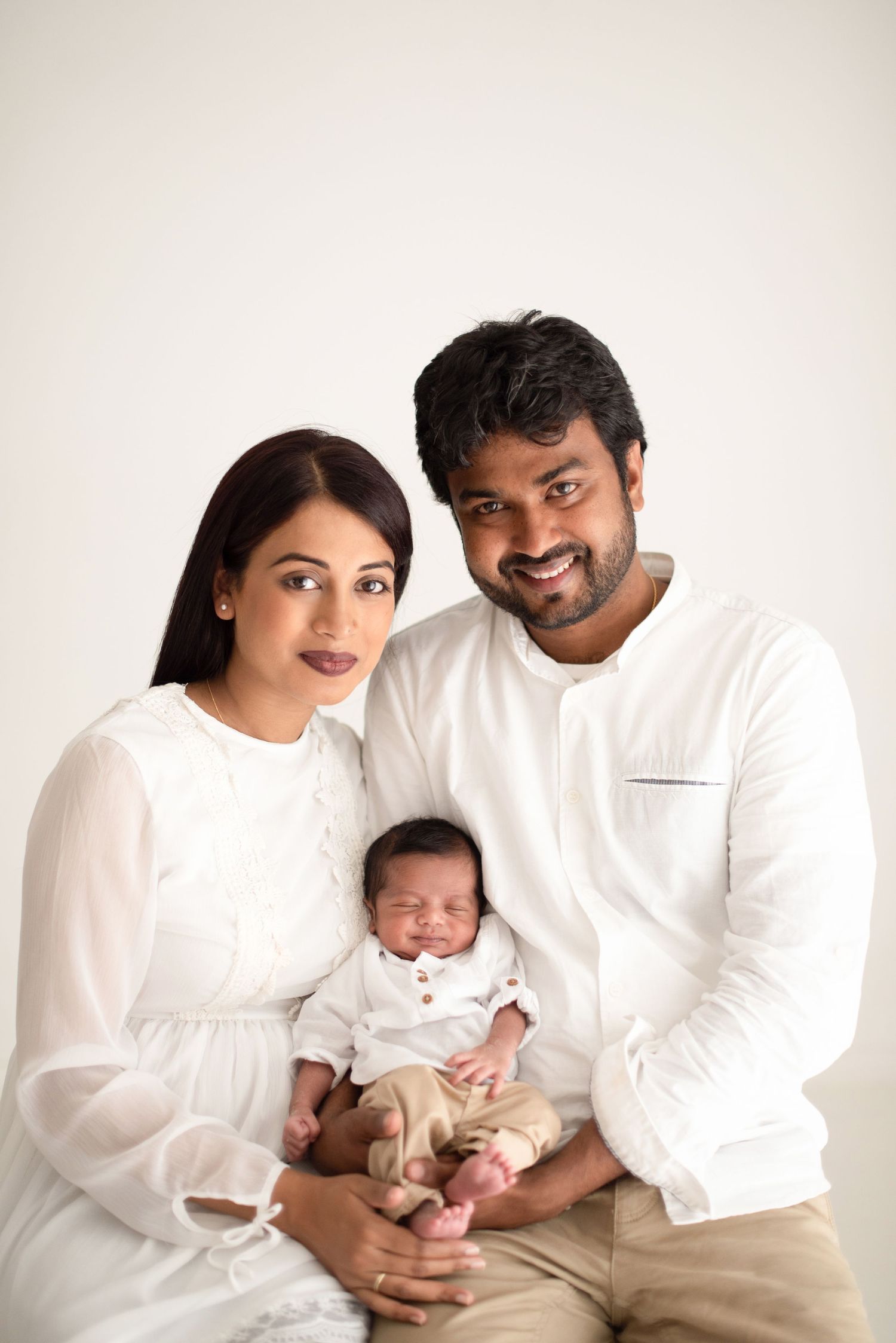 Family wearing white outfits poses together while holding their newborn baby in a studio portrait.