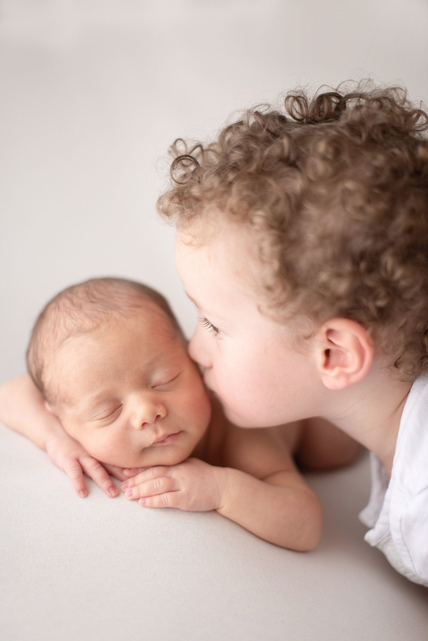A tender moment captured as a child with curly hair gently kisses a sleeping newborn baby.