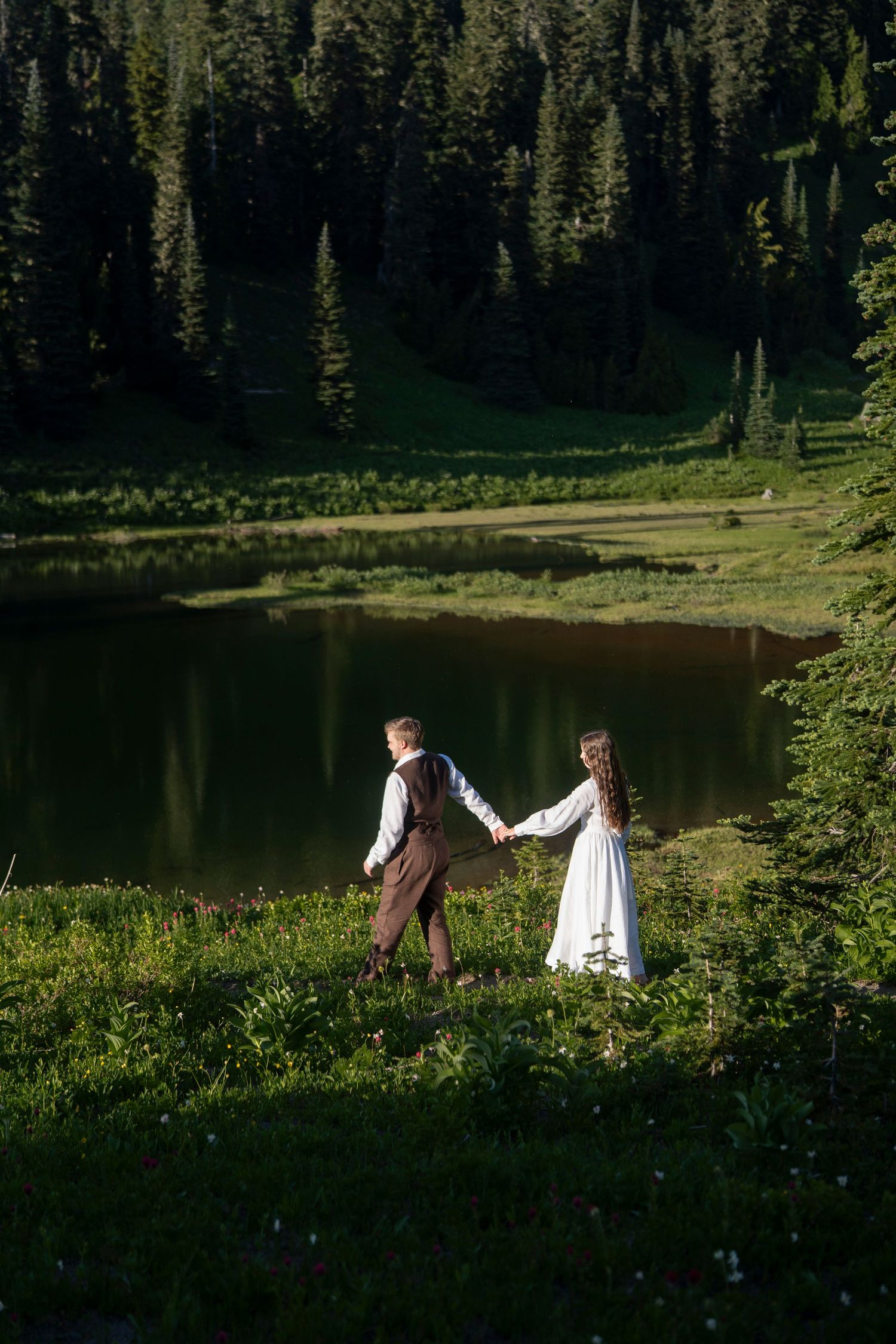 A couple sits by a serene mountain lake surrounded by evergreen forest on a sunny summer day.