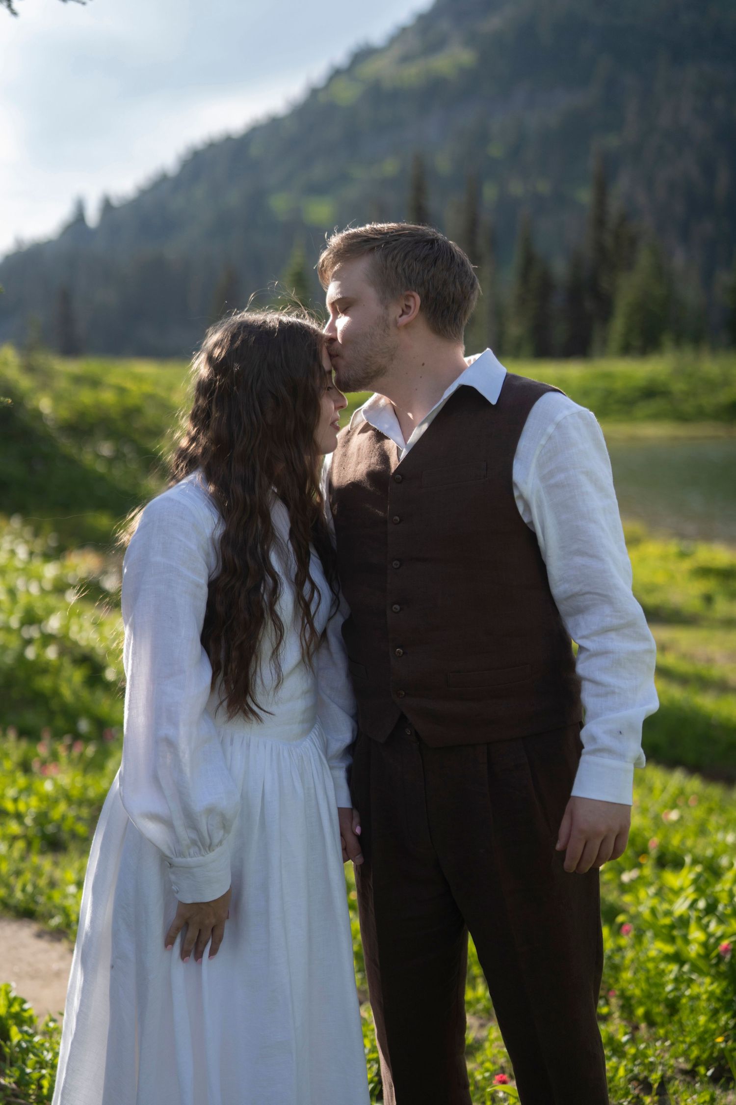 A romantic couple in wedding attire share a tender moment in a scenic mountain meadow with wildflowers and evergreens.