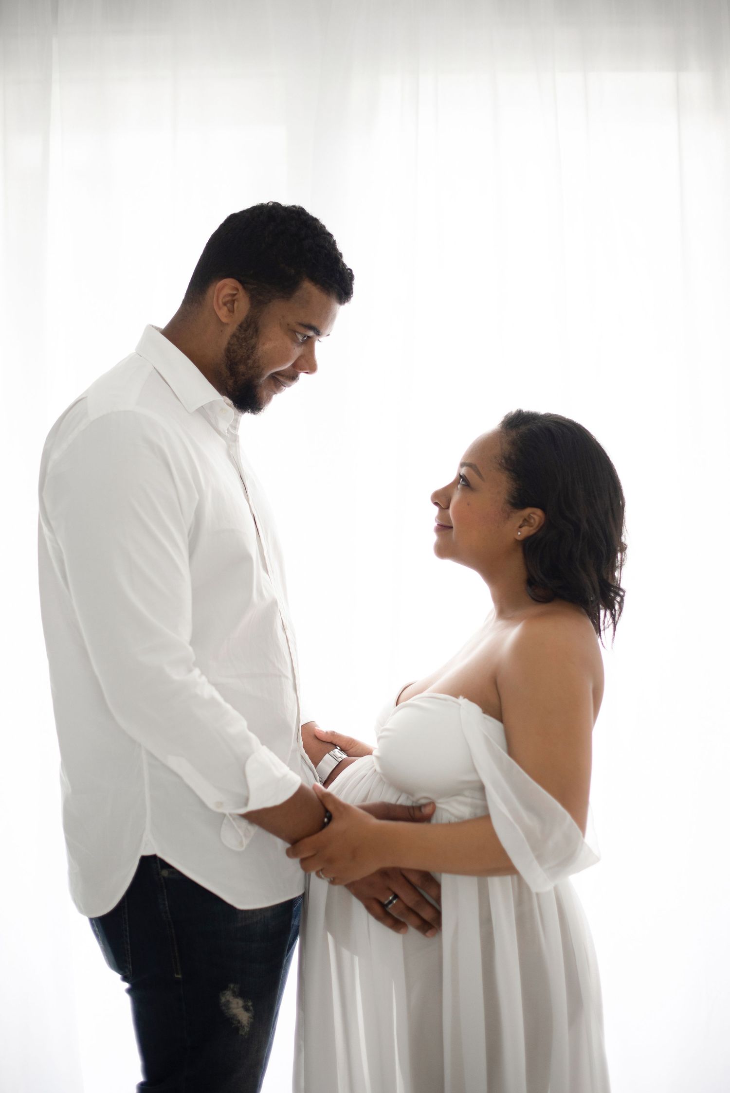 A couple in white clothing share an intimate moment during their maternity photoshoot.