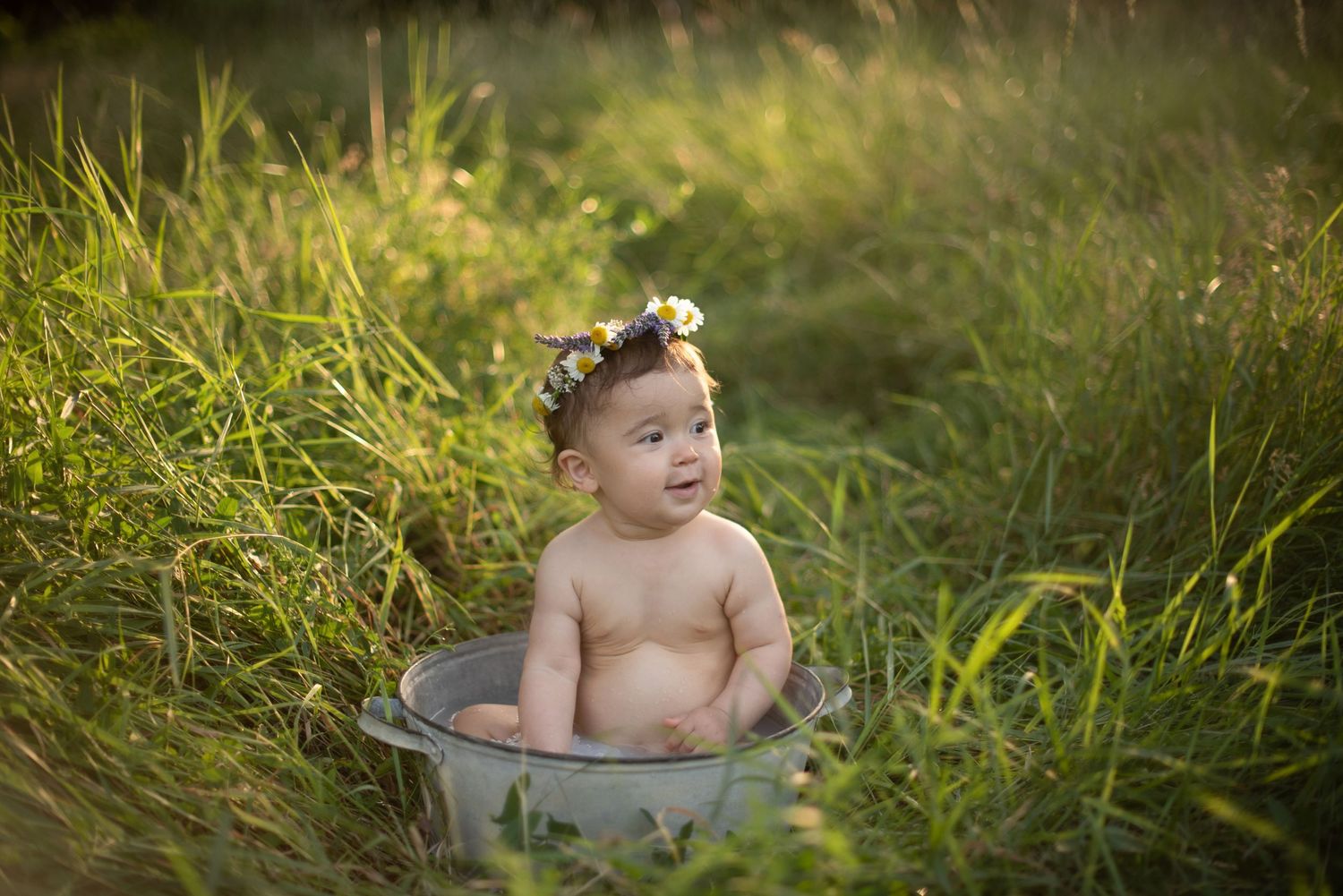 Baby with floral headpiece sits in galvanized tub surrounded by tall grass.