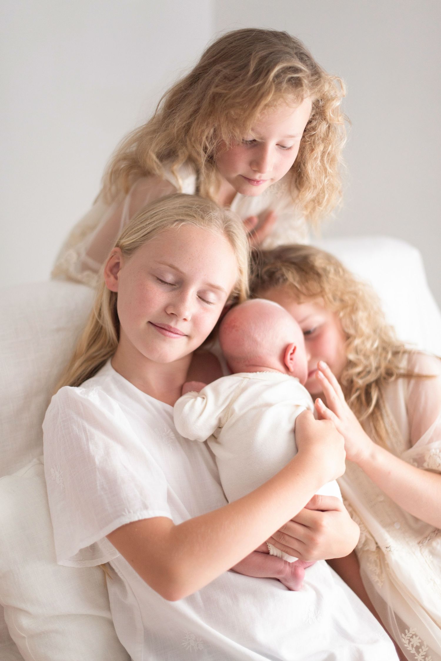 Siblings in white clothing sharing a tender moment with their newborn baby.