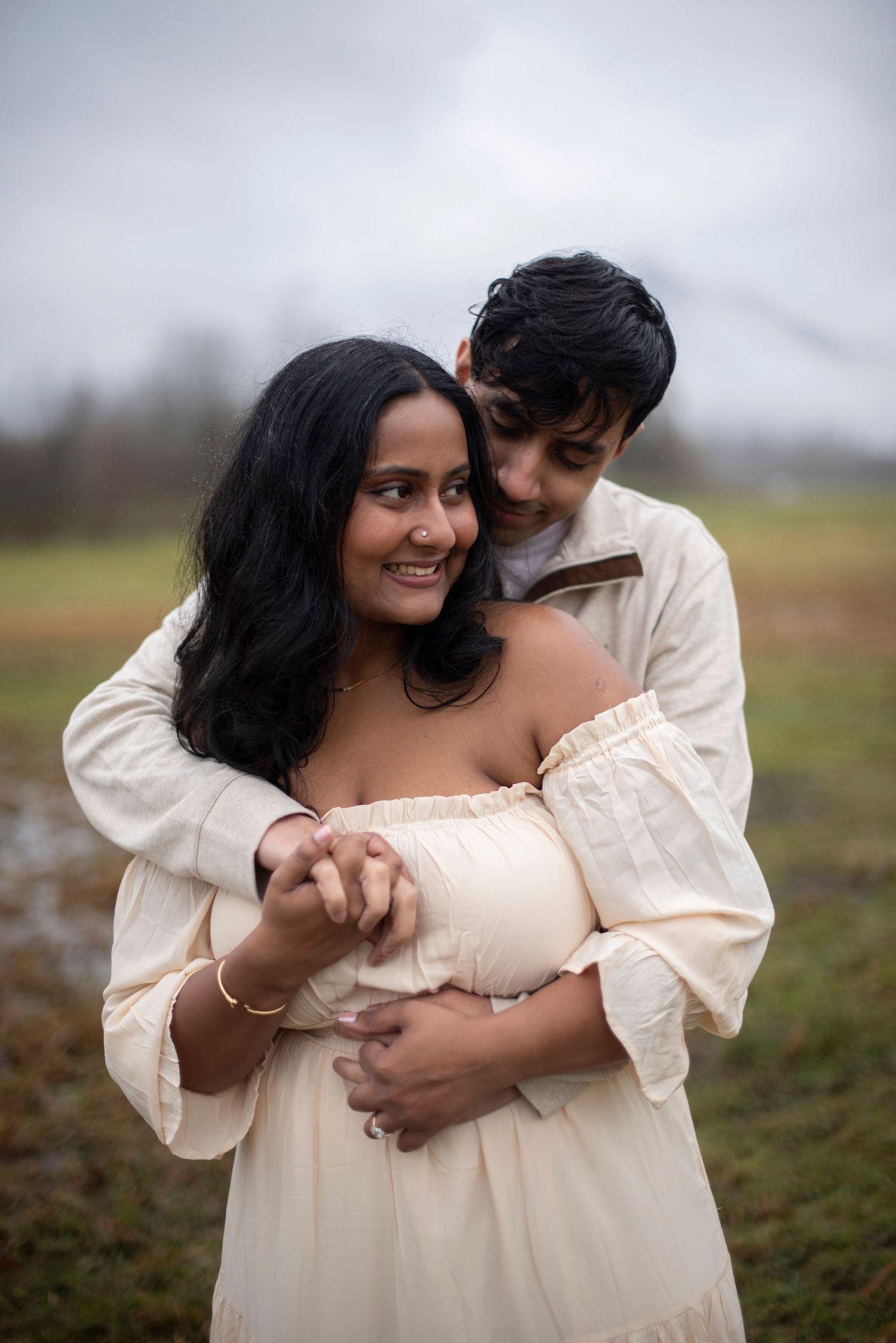 A romantic couple in cream-colored outfits embraces tenderly in a misty outdoor field setting.